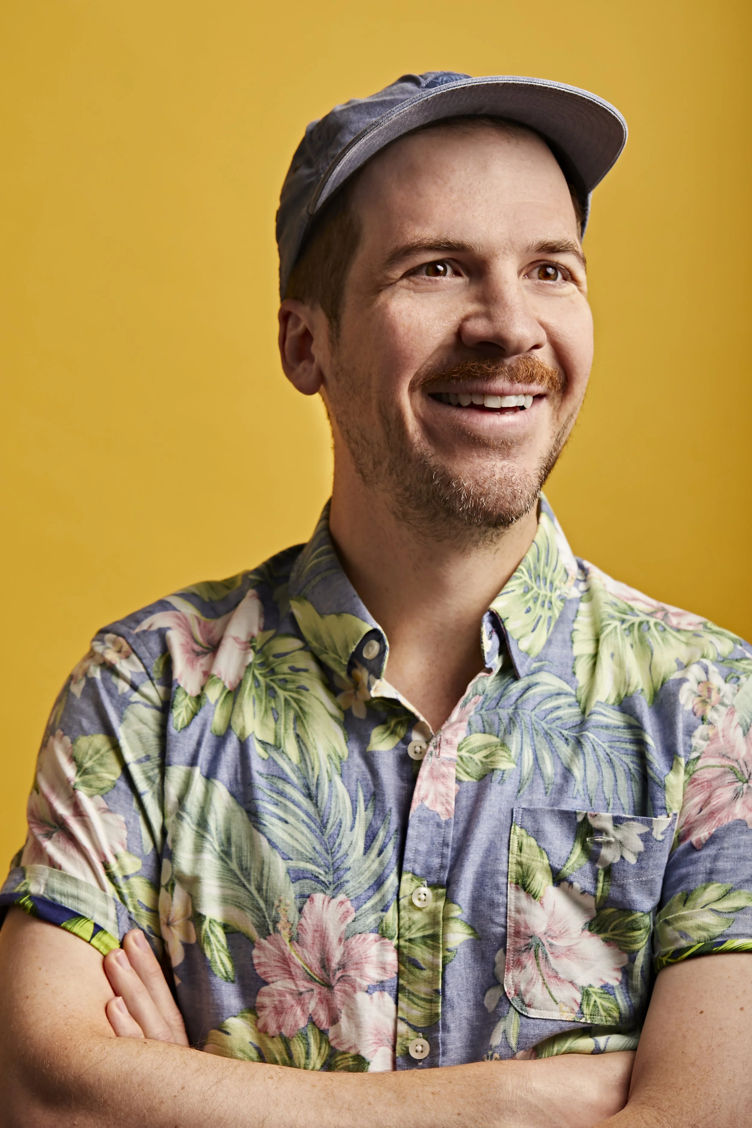 A smiling man with a short beard and mustache, wearing a gray cap and a colorful floral short-sleeve shirt against a yellow background.