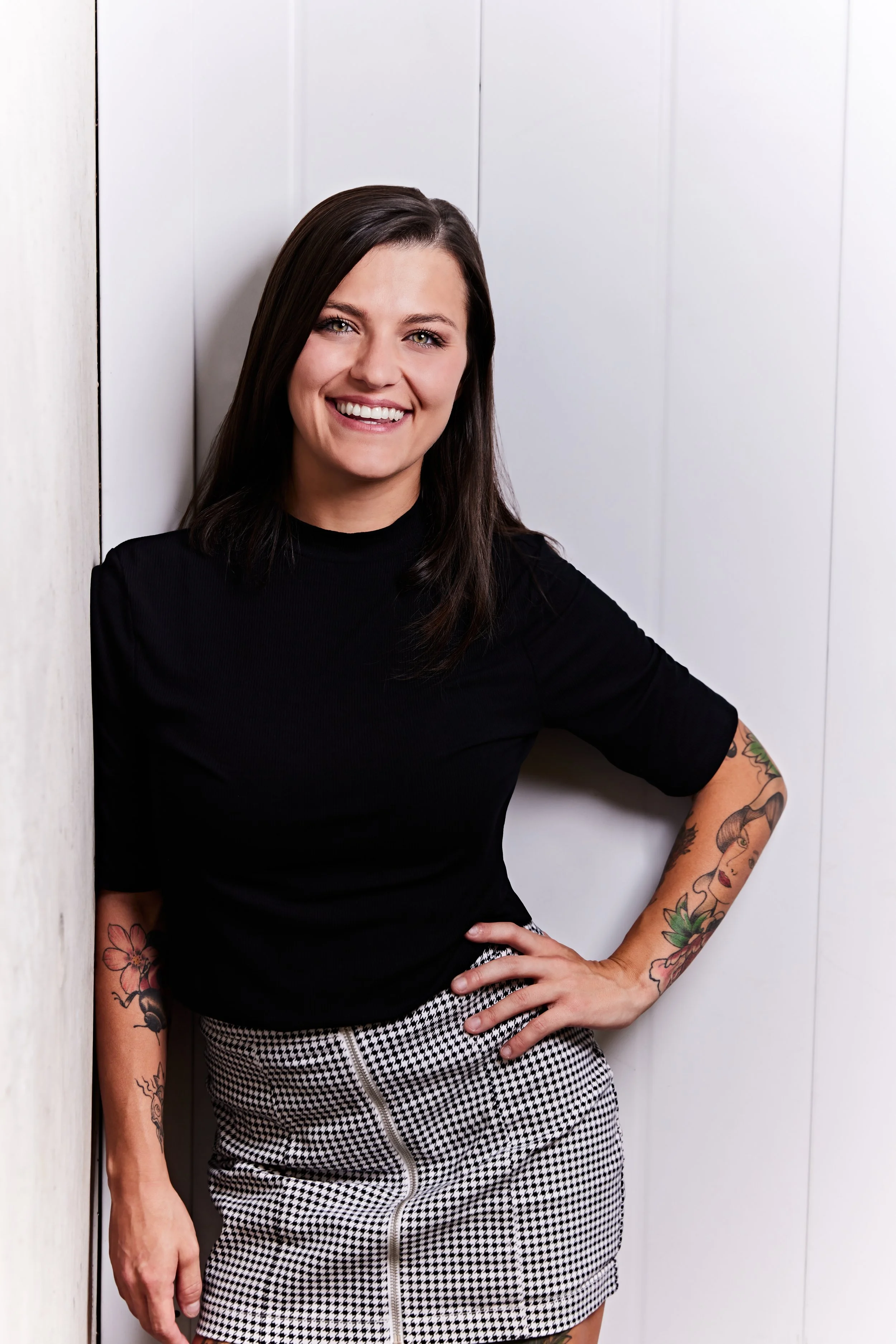 A woman with dark hair, wearing a black top and houndstooth skirt, smiling while leaning against a white wall.