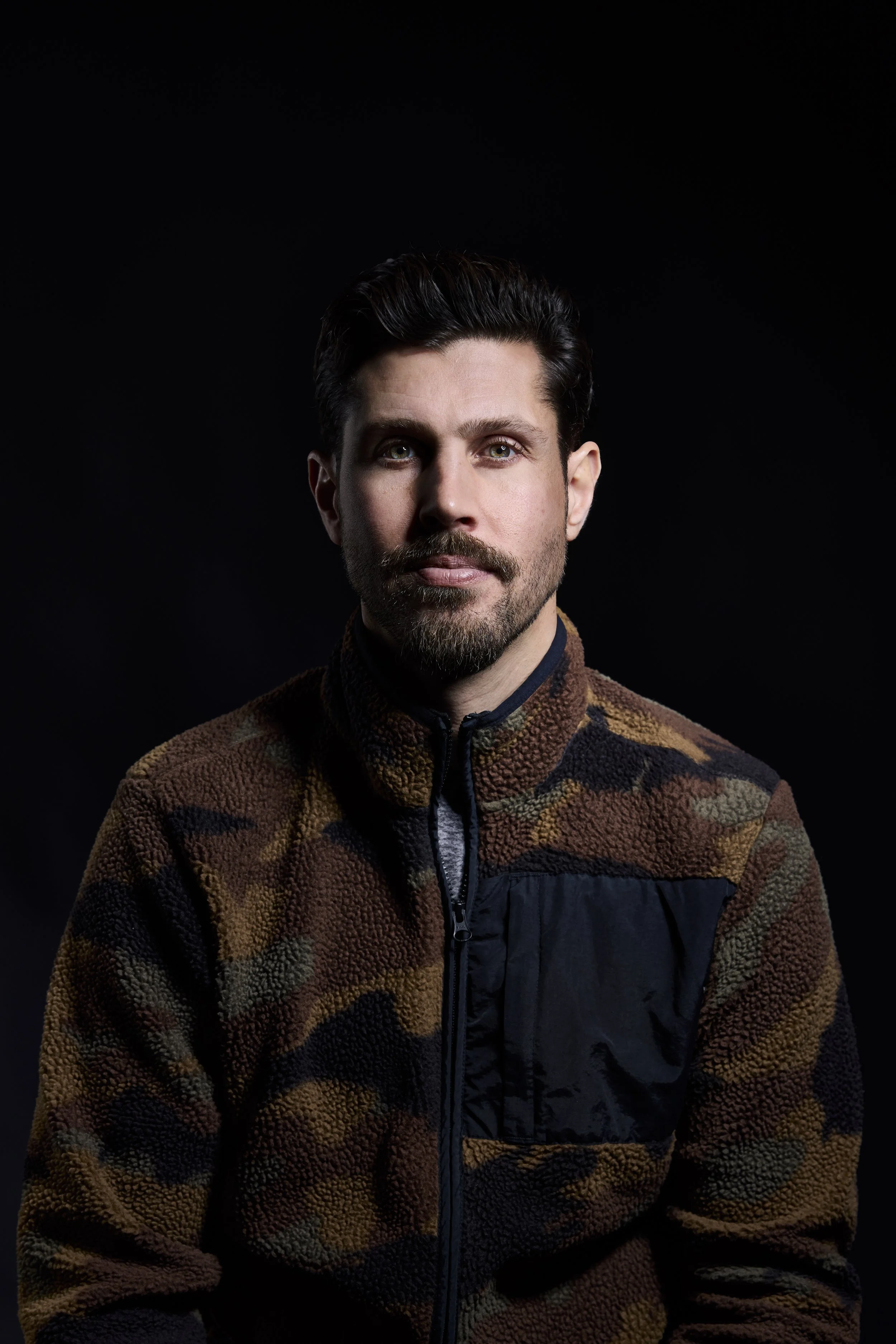 A man with dark hair and a beard, wearing a multicolored fleece jacket, poses against a black background.