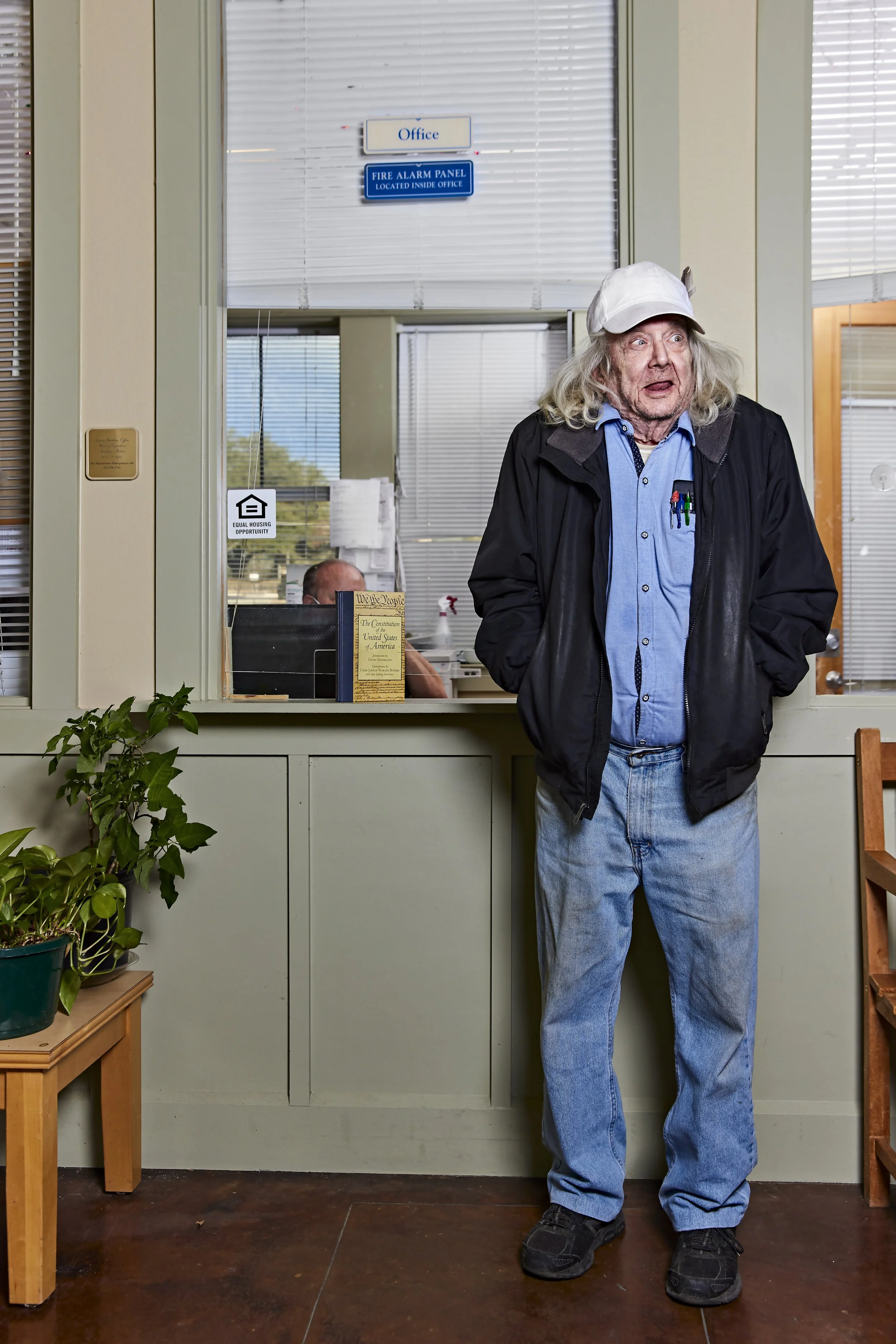 An elderly man with long white hair, wearing a white cap, blue shirt, black jacket, light blue jeans, and black shoes, stands in front of an office window with blinds. He has a surprised or confused expression on his face. Inside the office, a person