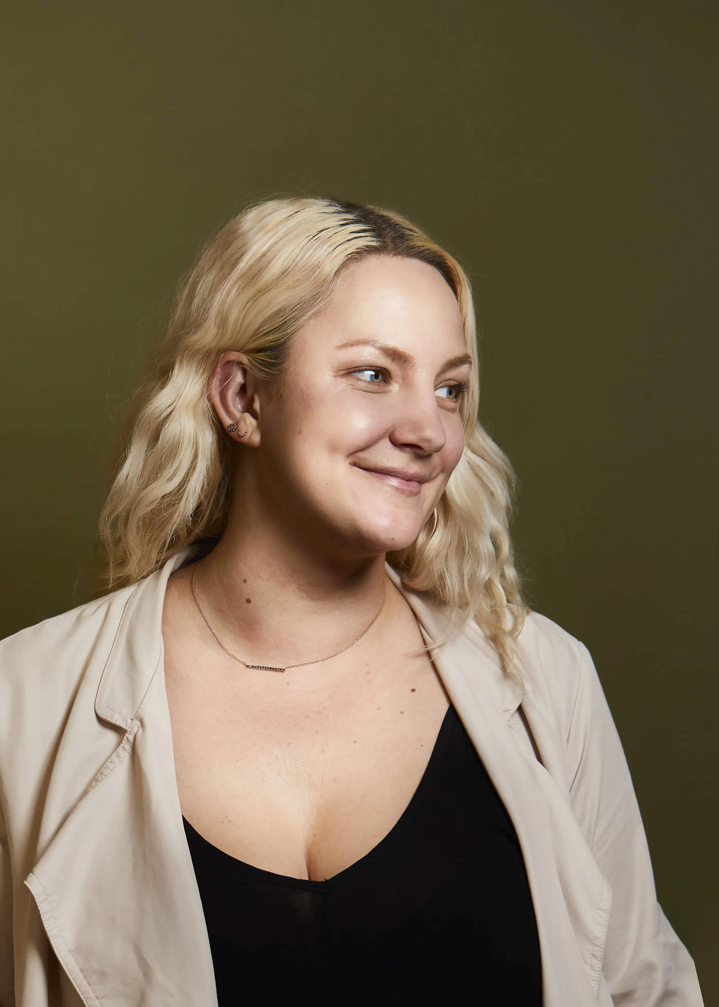 Portrait of a young woman with blonde curly hair, blue eyes, and light skin, smiling slightly, wearing a black top and a beige blazer, against a muted green background.