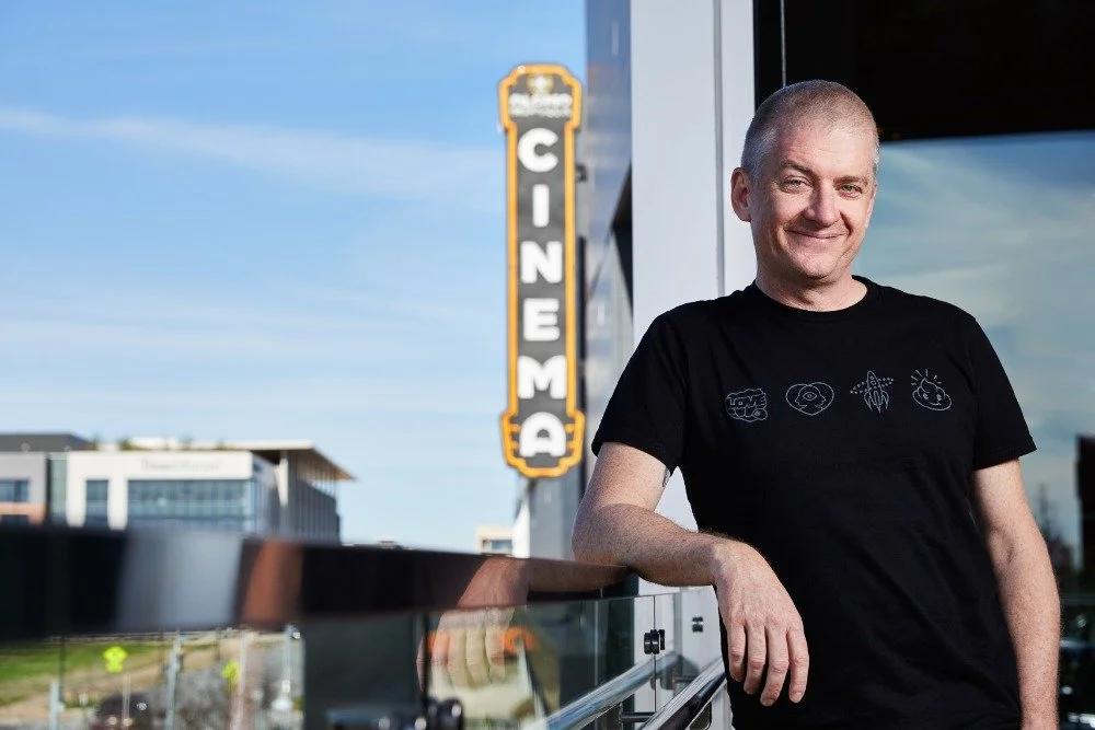A man with gray hair smiling, standing outside an American cinema theater, in front of a glass balcony railing with a blue sky background.
