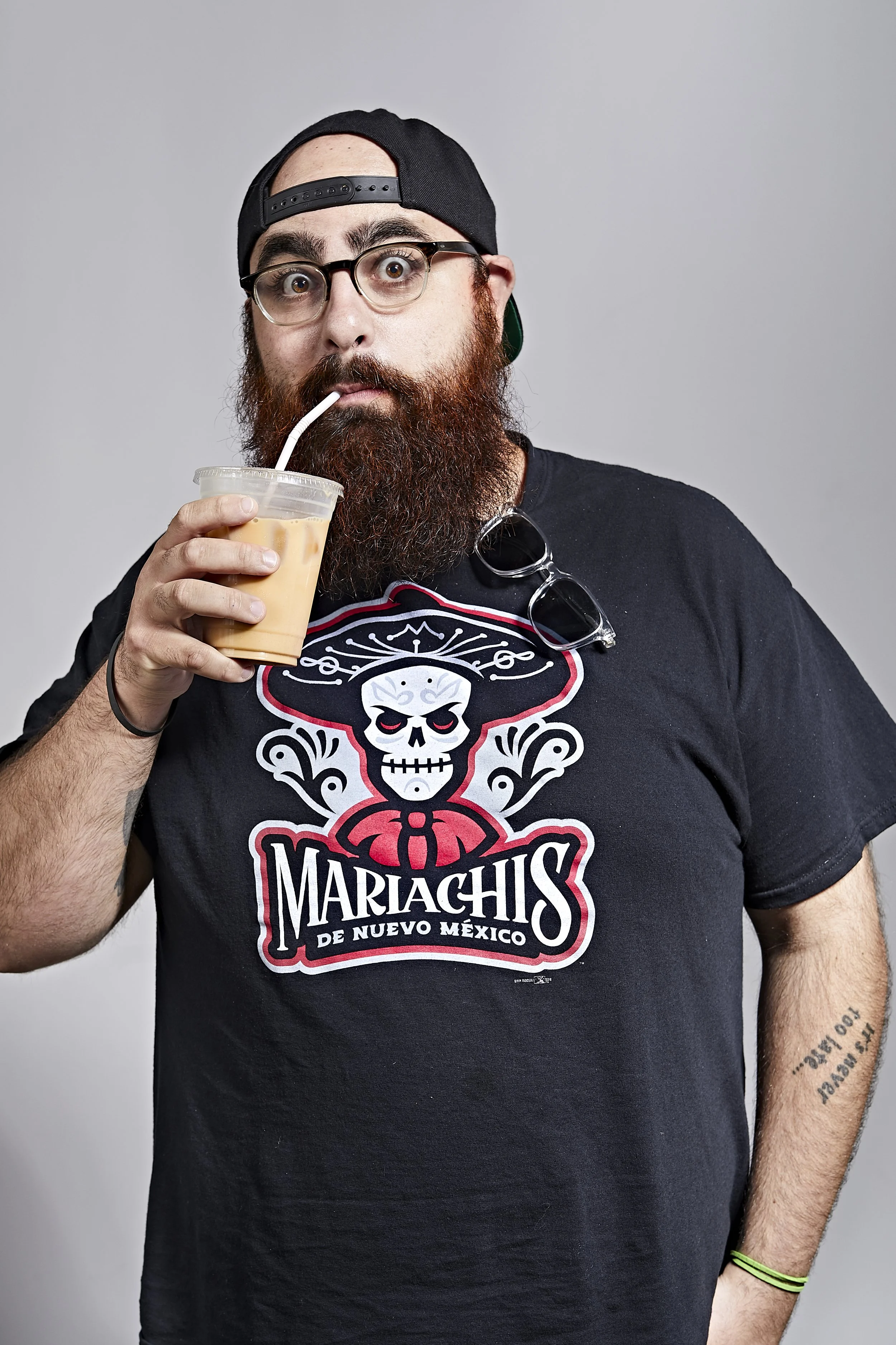 A bearded man wearing glasses, a black baseball cap backwards, and a black T-shirt with a skull and mariachis graphic, drinking iced coffee with a straw.