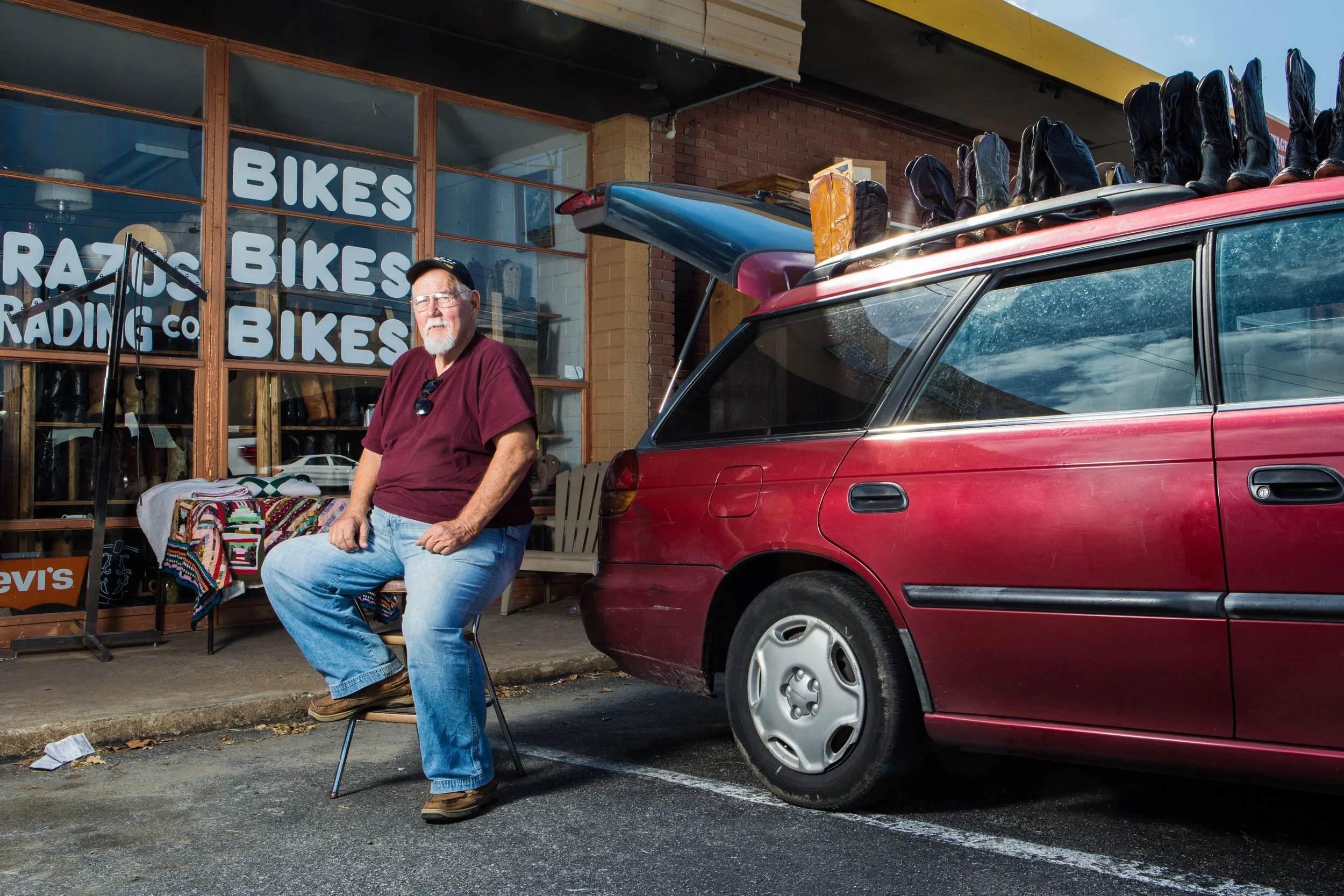 A man sitting on a chair outside a bike shop with large windows displaying the word "BIKES" multiple times, a red car parked nearby with surfboards and boots on the roof, and various items displayed in front of the store.