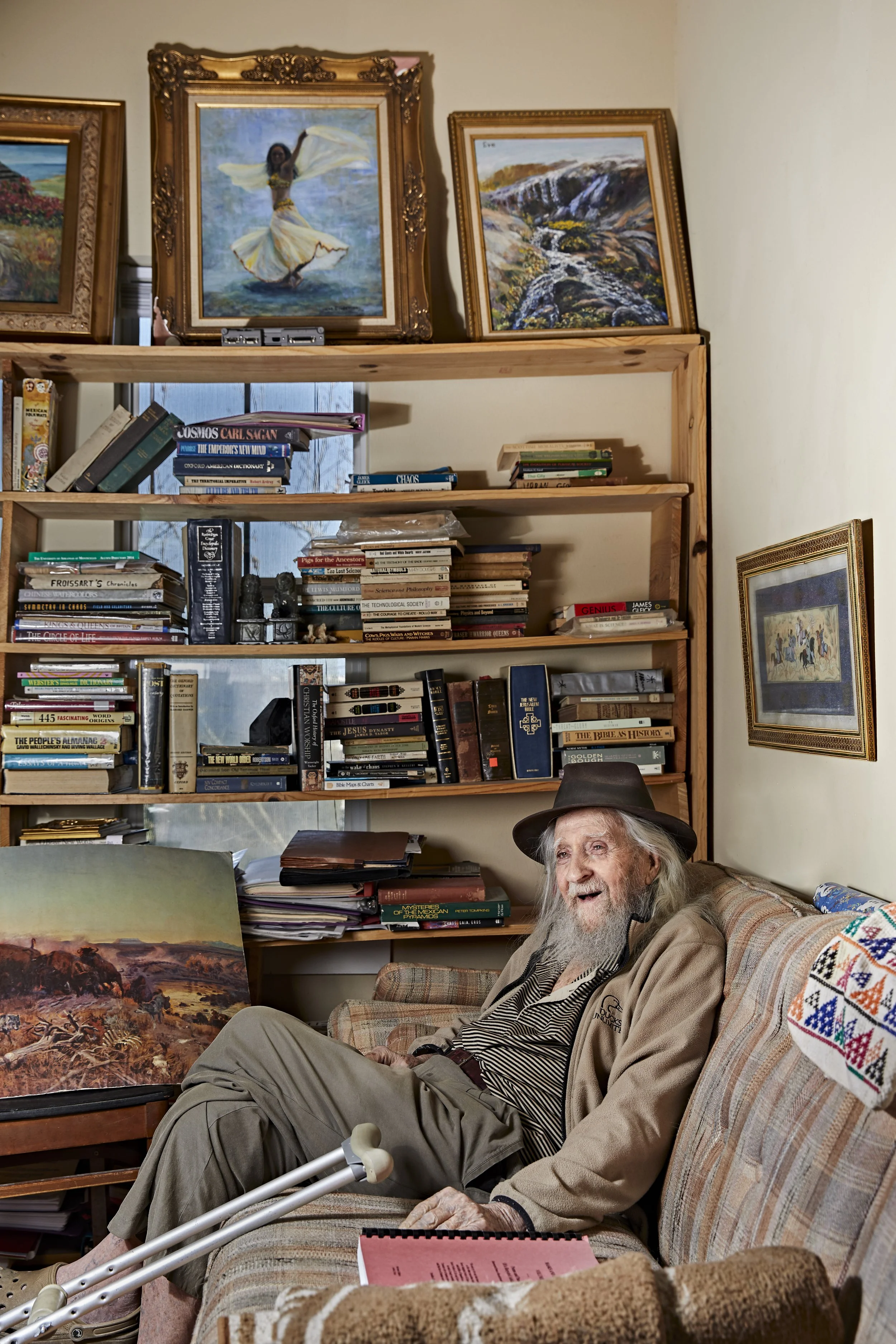An elderly man with long gray hair and beard, wearing a fedora hat, striped shirt, beige jacket, and tan pants, sitting on a patterned couch in a room filled with books, paintings, and art supplies. He is smiling and appears to be engaged in conversa