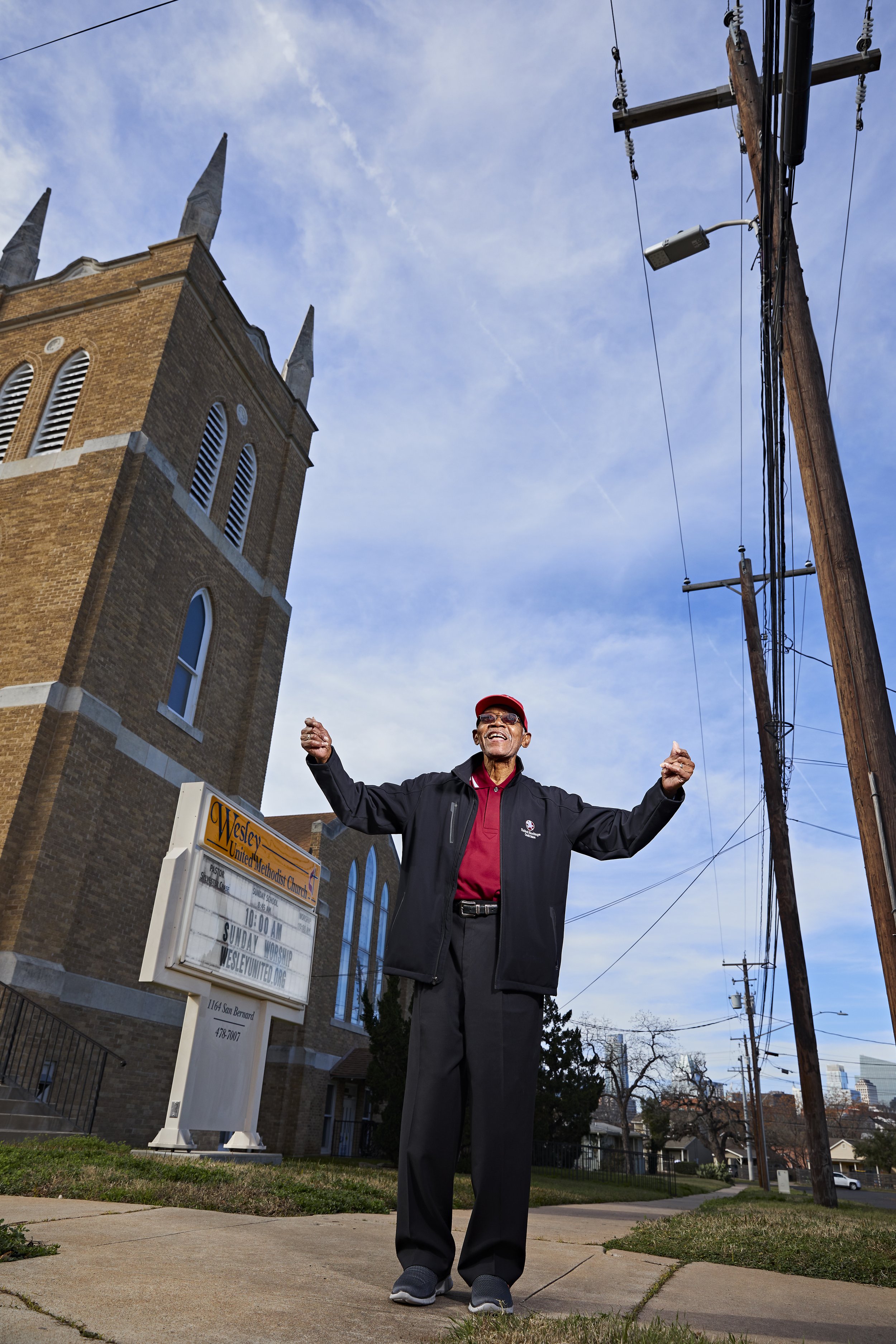 An elderly man standing on a sidewalk with his arms raised in front of a church and power lines on a clear day.