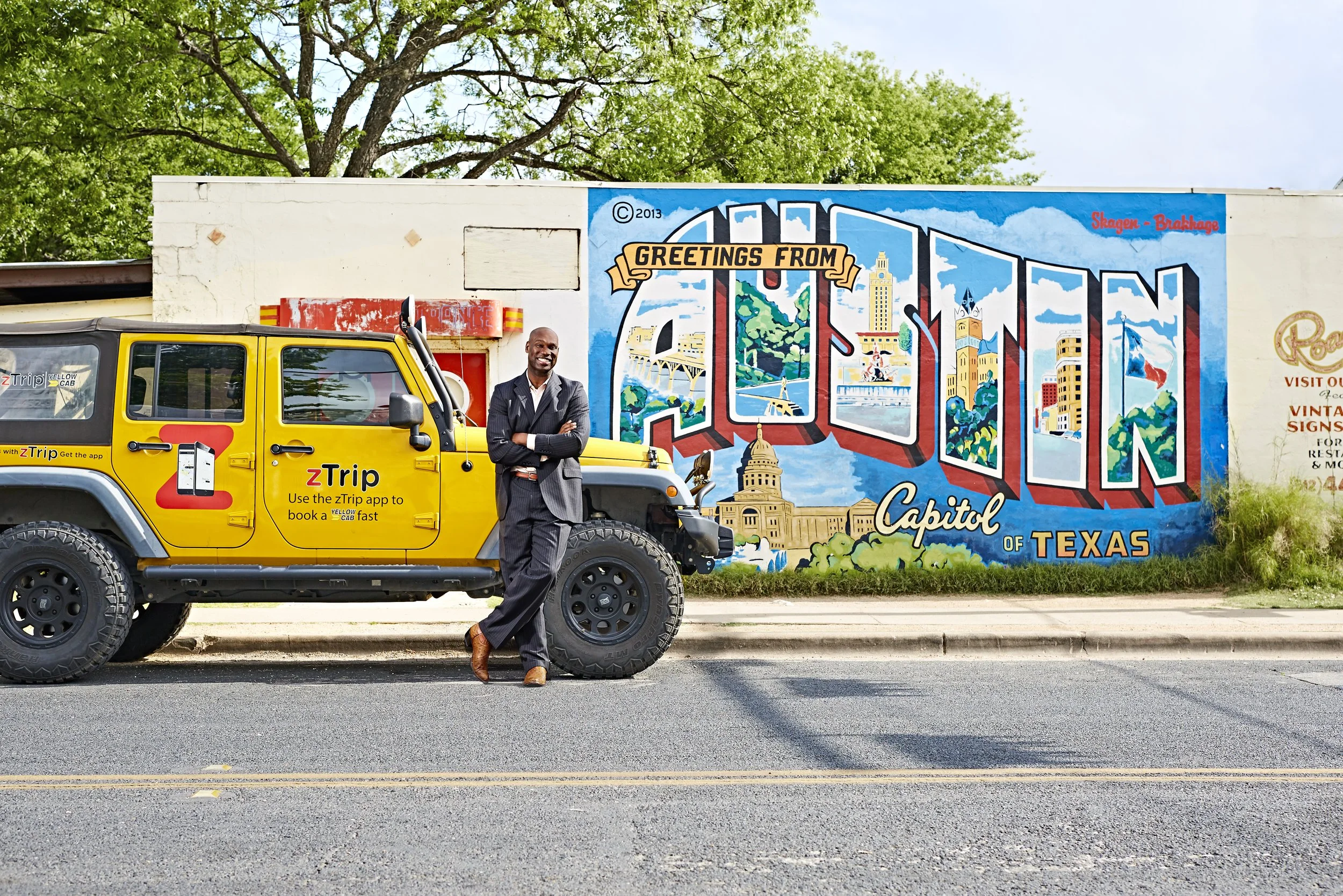 A man in a suit leaning against a yellow taxi with ZTrip logo on a city street in front of a colorful mural that reads "Greetings from Austin" with images of landmarks of Austin, Texas.