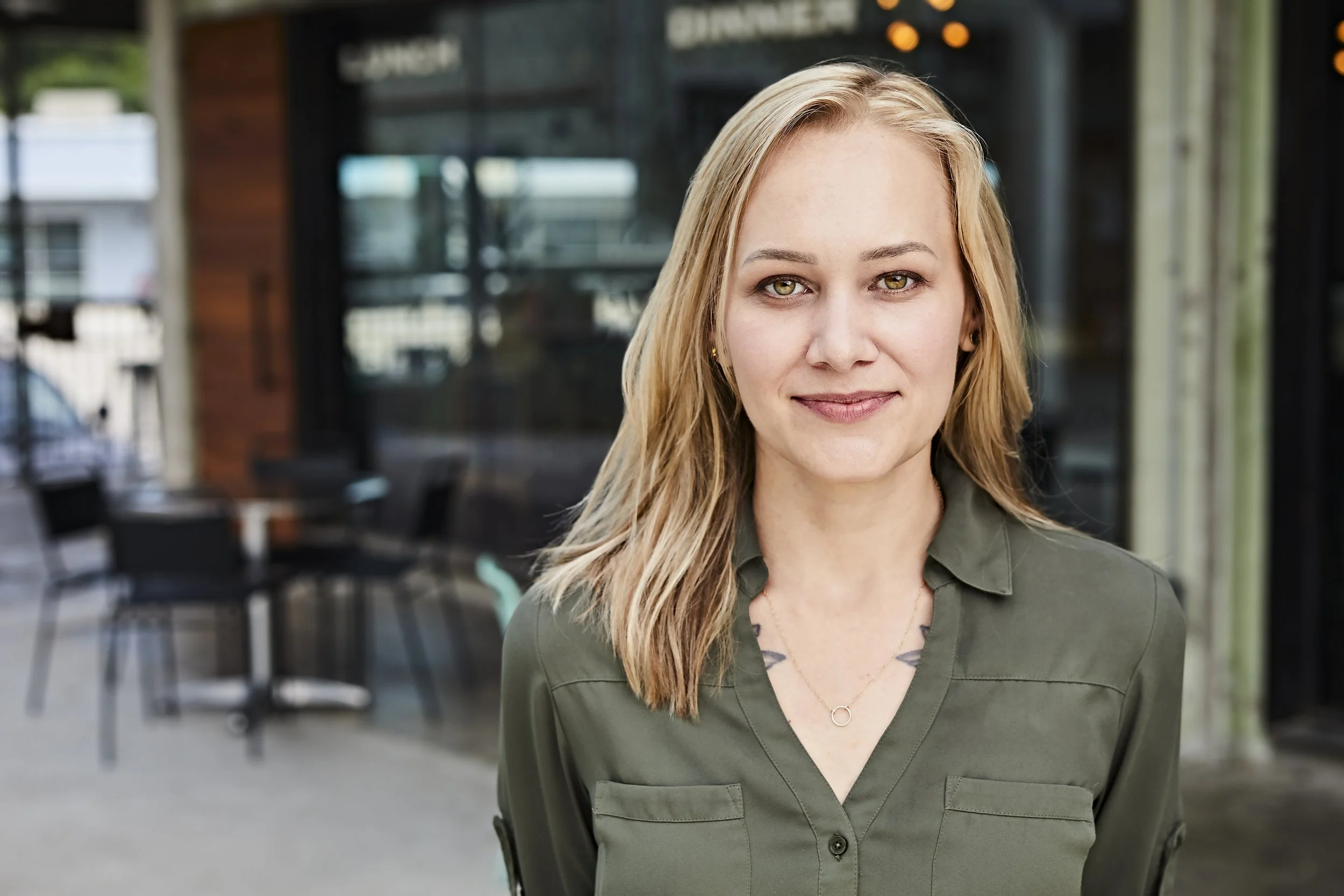 A woman with blonde hair and green eyes standing outdoors, smiling, with a modern building with glass windows behind her.