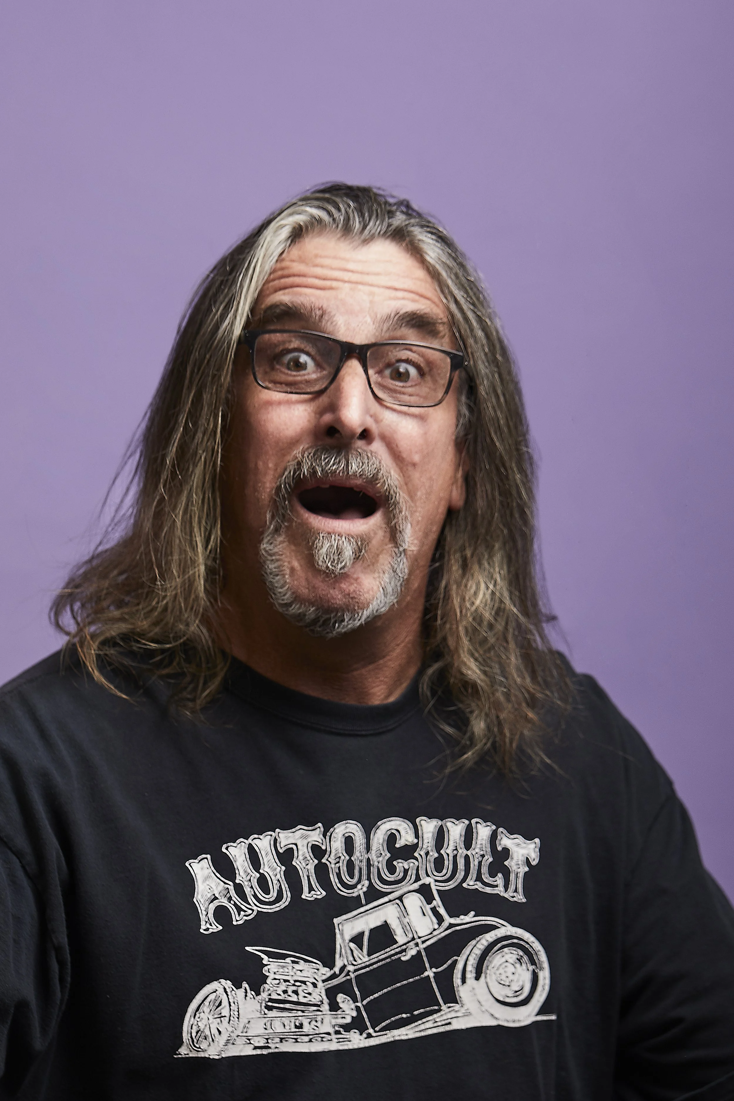 A man with long hair, glasses, and a beard has an surprised or excited expression, wearing a black T-shirt with a graphic design of a vintage car and the word 'AUTOCULT' on it, against a purple background.