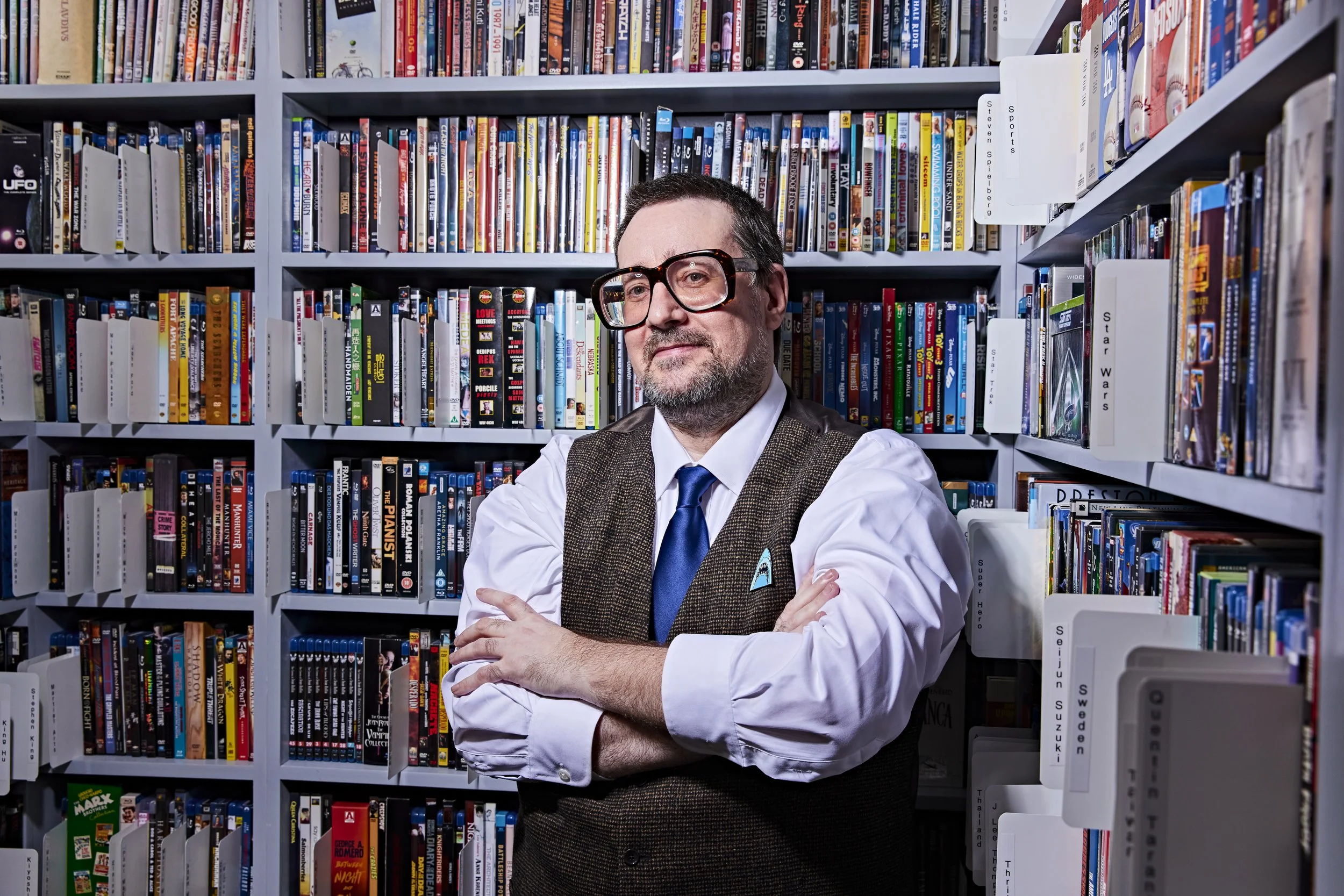 A man with glasses, wearing a white shirt, blue tie, and brown vest, standing in front of bookshelves filled with comics and books, with his arms crossed.