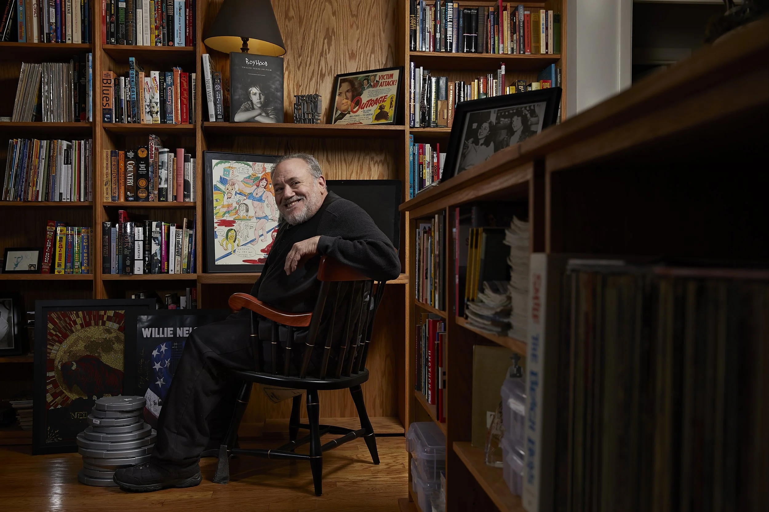 A smiling man sitting on a wooden chair in a room with wooden bookshelves filled with books, posters, and framed artwork.