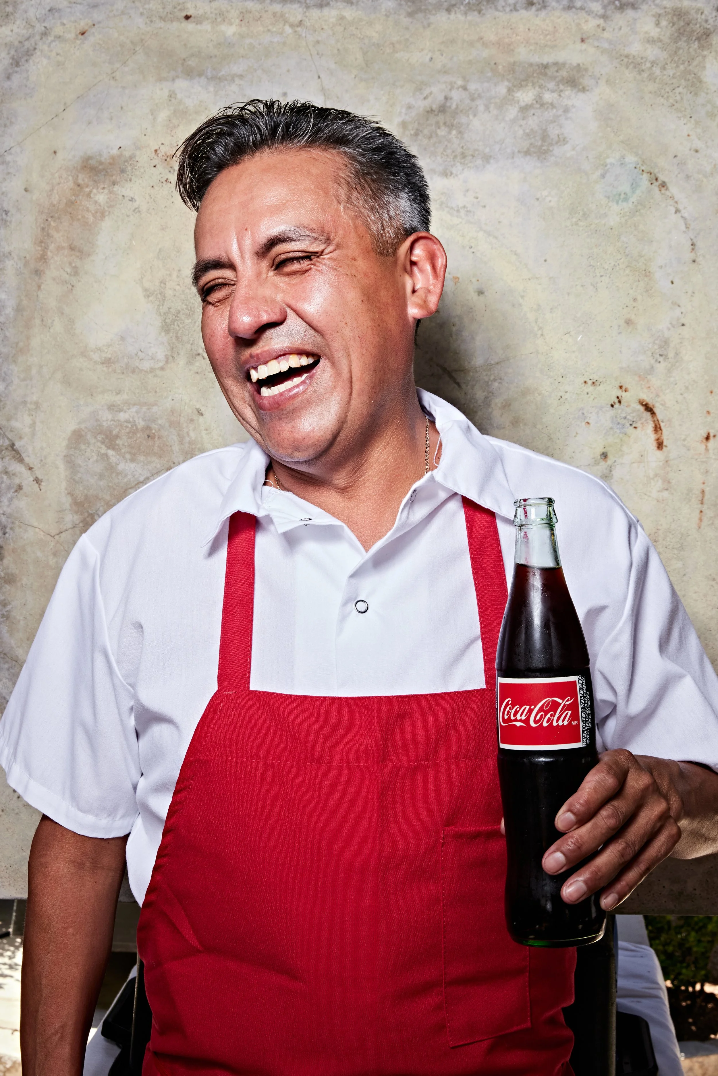 A man wearing a white shirt and red apron, laughing and holding a bottle of Coca-Cola, standing against a textured wall.