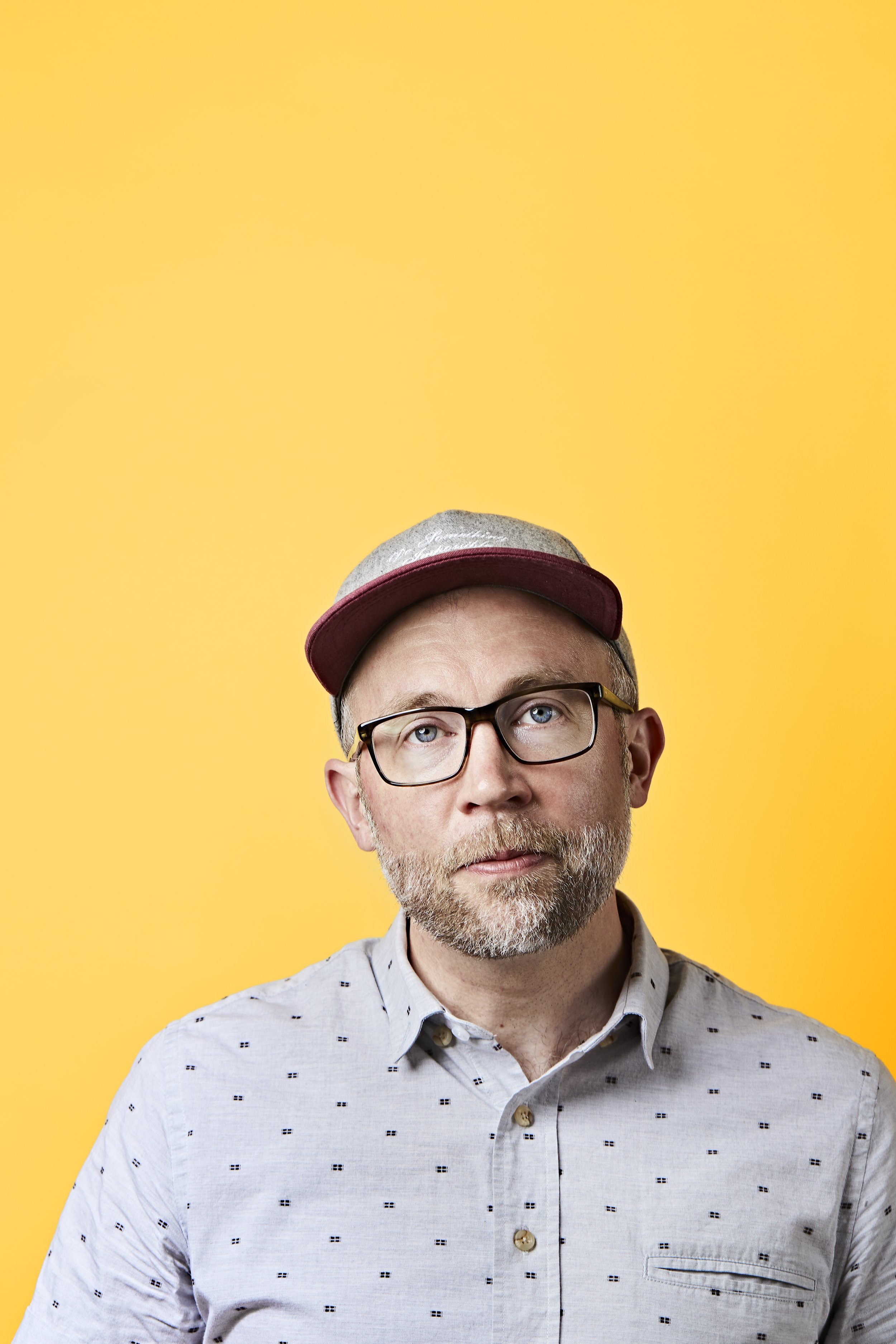 A man with glasses, a beard, and a gray and burgundy baseball cap, wearing a light gray shirt with small dark patterns, standing against a yellow background.
