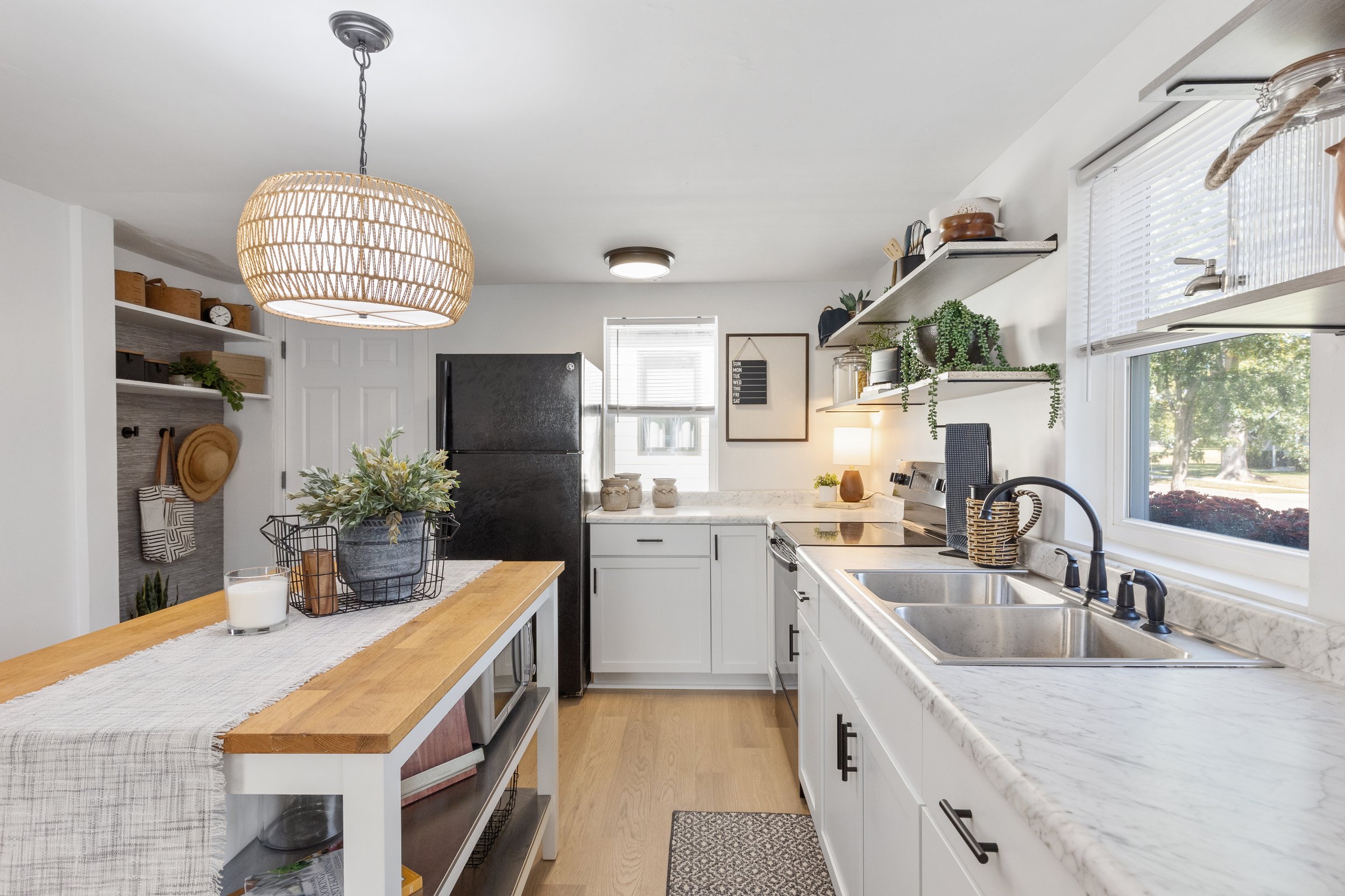 Kitchen with white cabinets, marble countertop, black refrigerator, open shelves with plants and dishes, window with blinds, wooden island with decorative plant, candle, and runner, hardwood floor, and pendant light.
