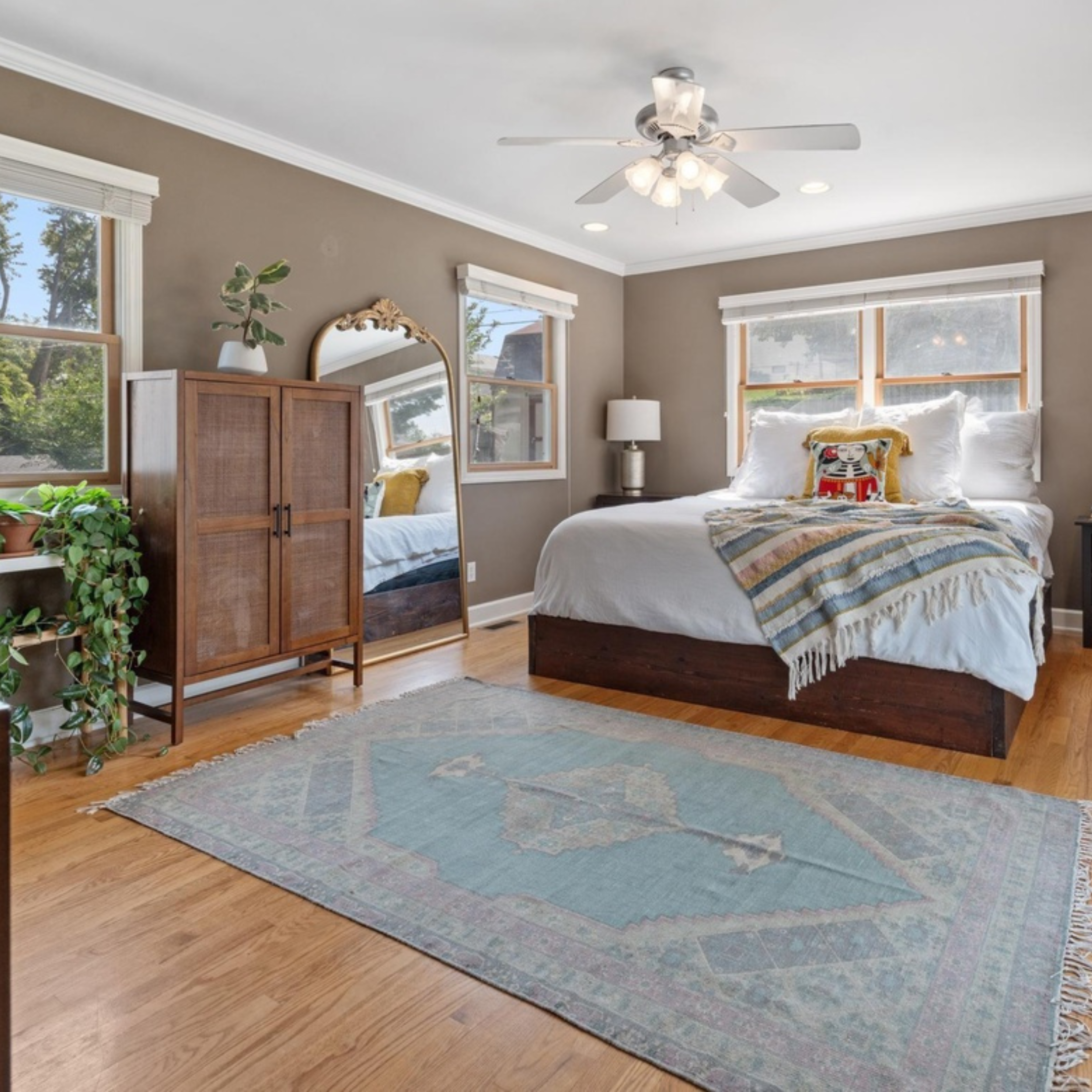 A bedroom with a bed, wooden floor, and multiple windows. There is a wooden wardrobe with a potted plant on top, a large mirror leaning against the wall, a small nightstand with a lamp, and a rug on the floor.