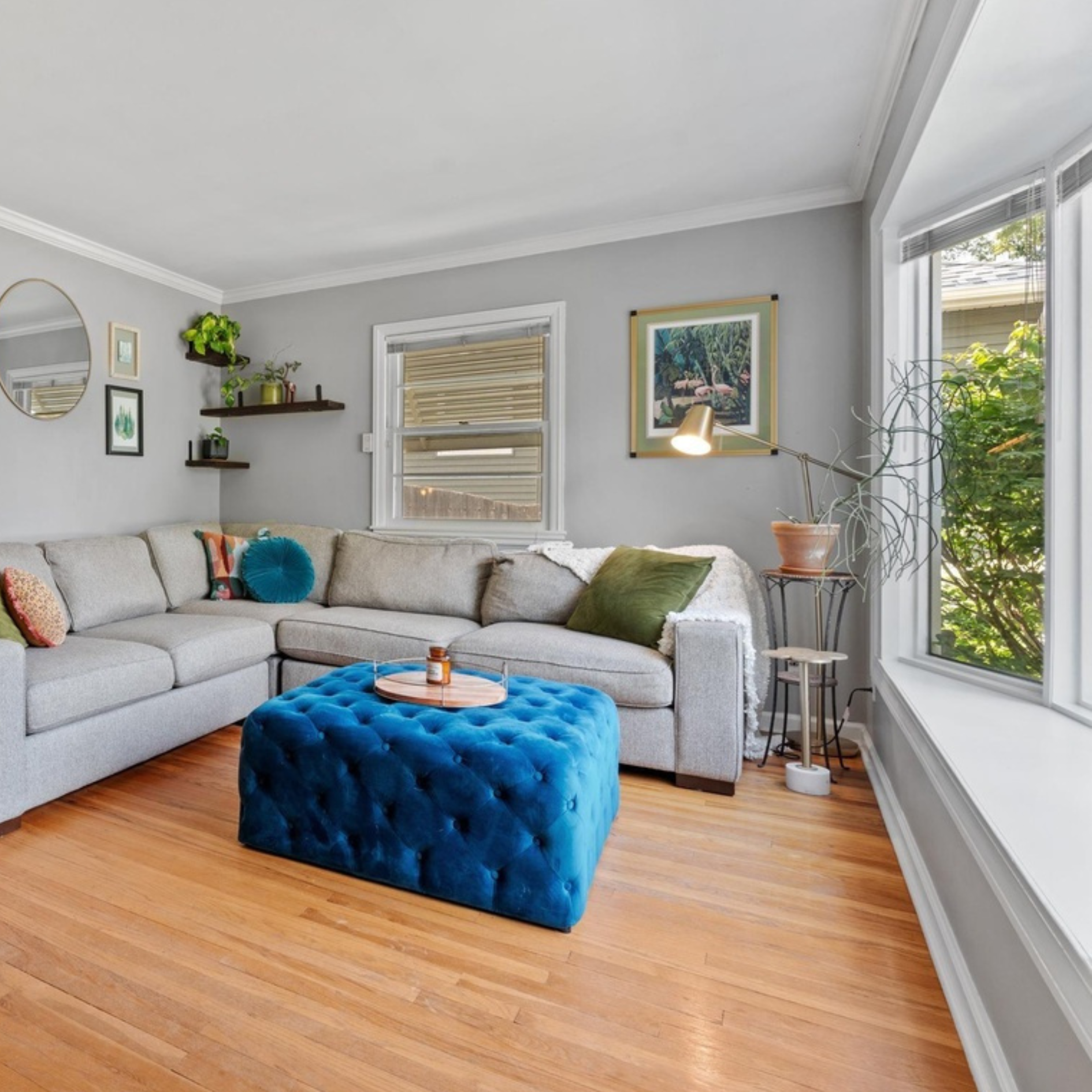 Living room with a light-colored sectional sofa, colorful cushions, a blue tufted ottoman with a tray and bottle, wooden floor, and large windows with greenery outside.