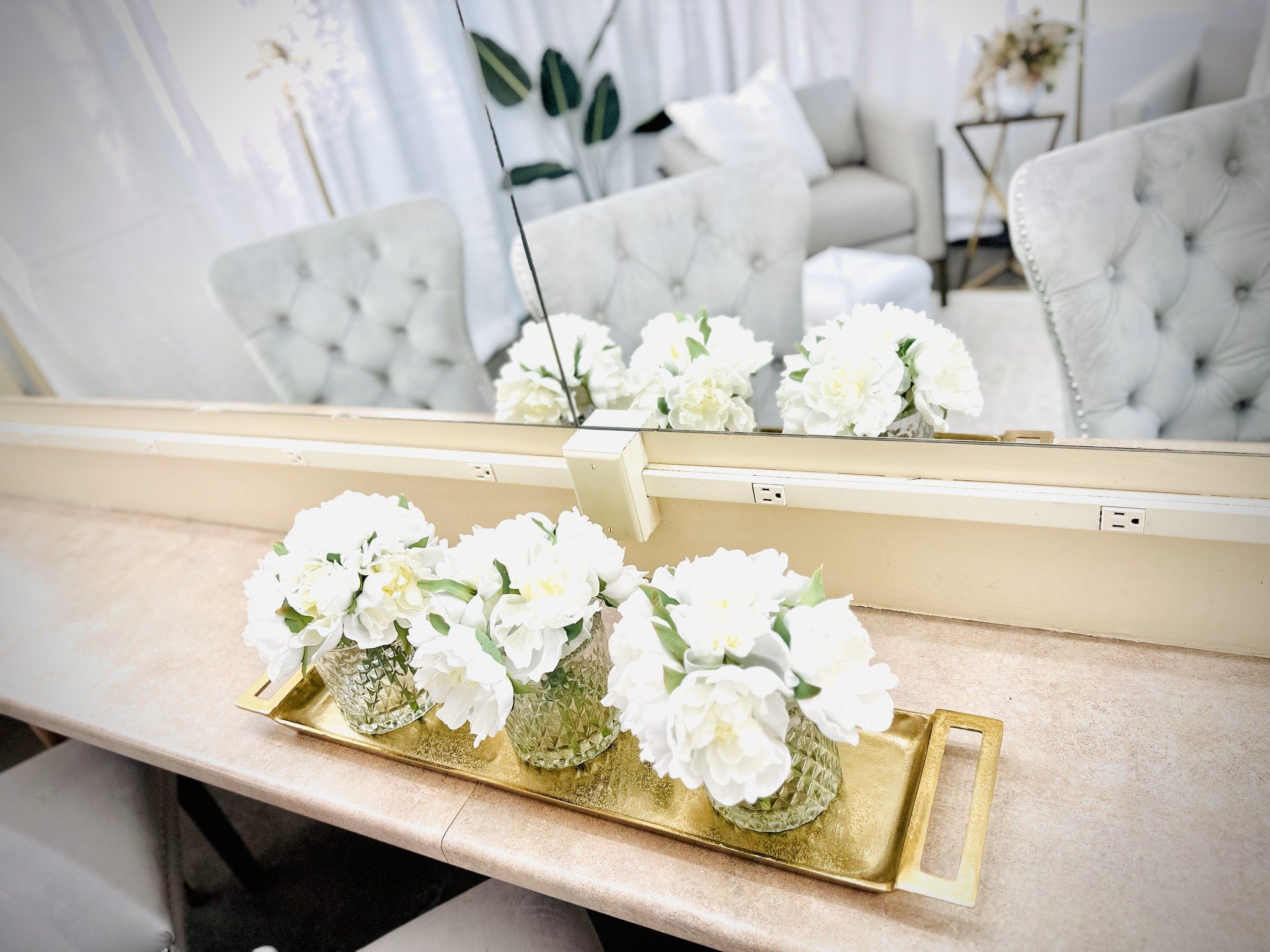 White artificial flowers in textured glass vases on a gold tray in front of a mirror, with a living room featuring tufted armchairs, a white sofa, and side tables in the background.