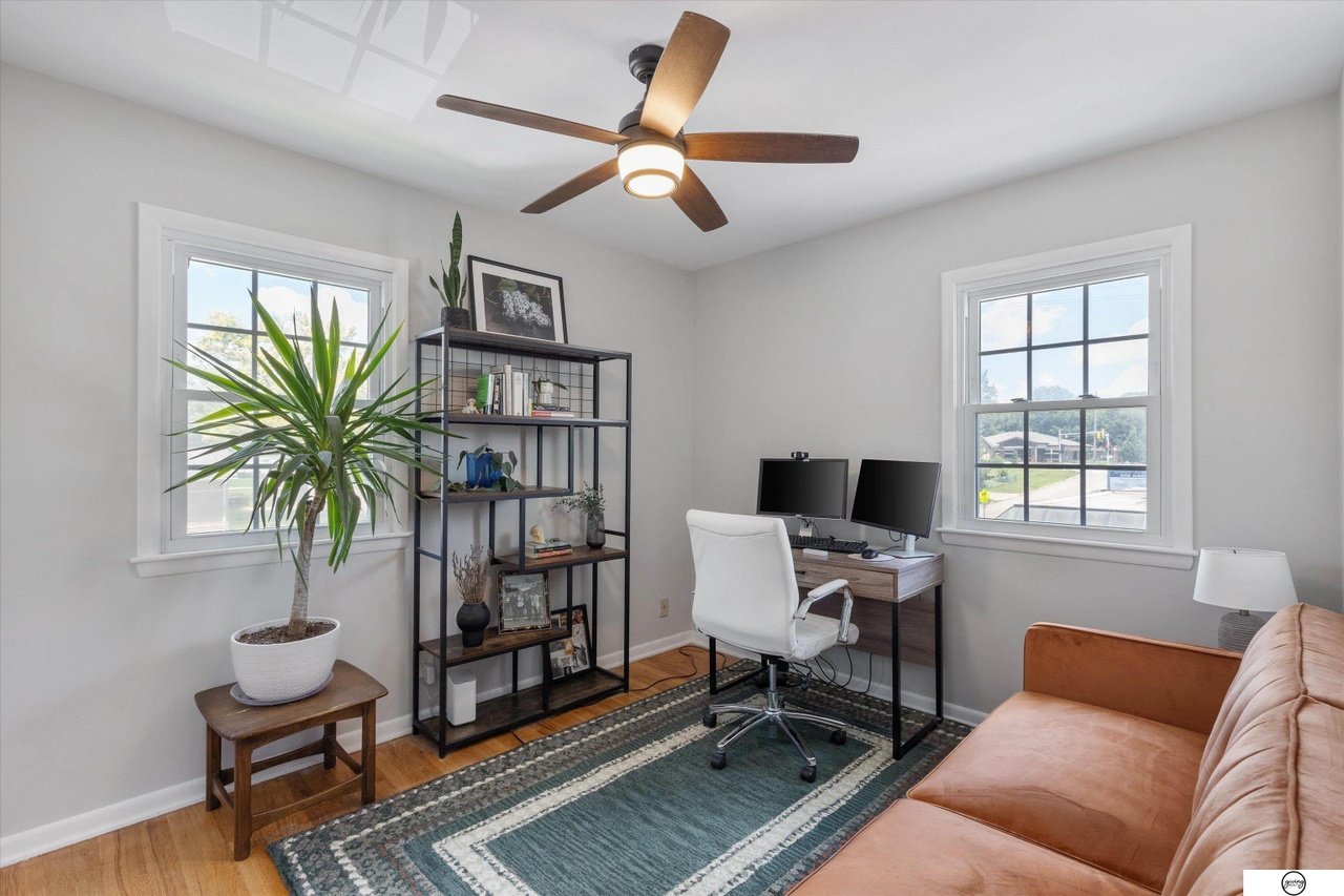Home office with white walls, two windows, a ceiling fan, a potted plant, a metal shelf with books and decor, a desk with dual monitors, a white office chair, a tan sofa, a patterned rug, and a small side table with a lamp.
