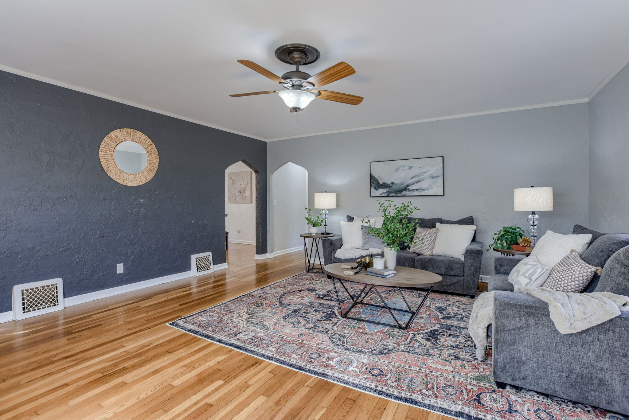 Living room with dark gray and light gray walls, hardwood floors, a ceiling fan, arranged with gray sofas, a round wooden coffee table, table lamps, an area rug, and wall art.