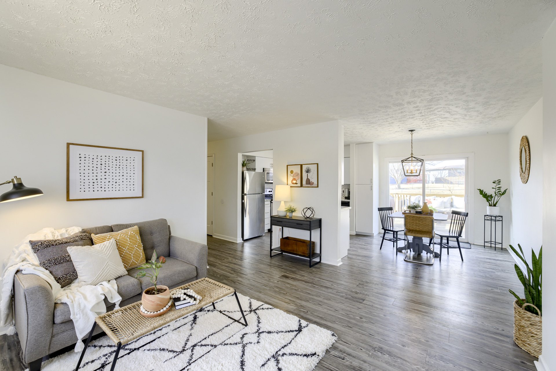 Living room and dining area with white walls, gray floor, and modern decor, including a gray couch with pillows, a coffee table, a small console table with a lamp and decorative items, and a round dining table with chairs near sliding glass door lett