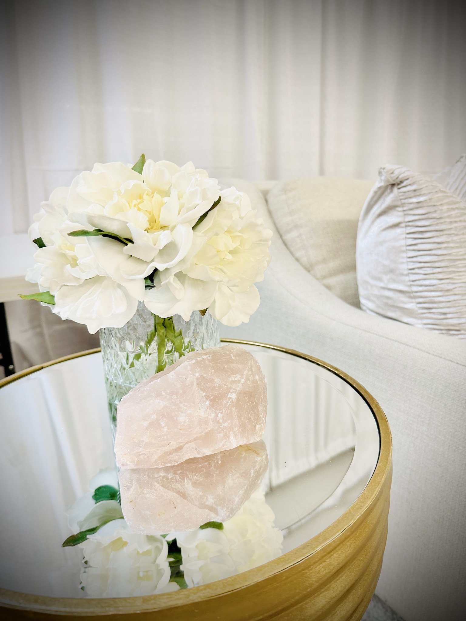 A glass-topped table with a large white peony flower in a textured glass vase, a pink Himalayan salt rock, and white flowers reflected in the table's mirror surface. A beige upholstered sofa with textured and striped pillows is in the background.