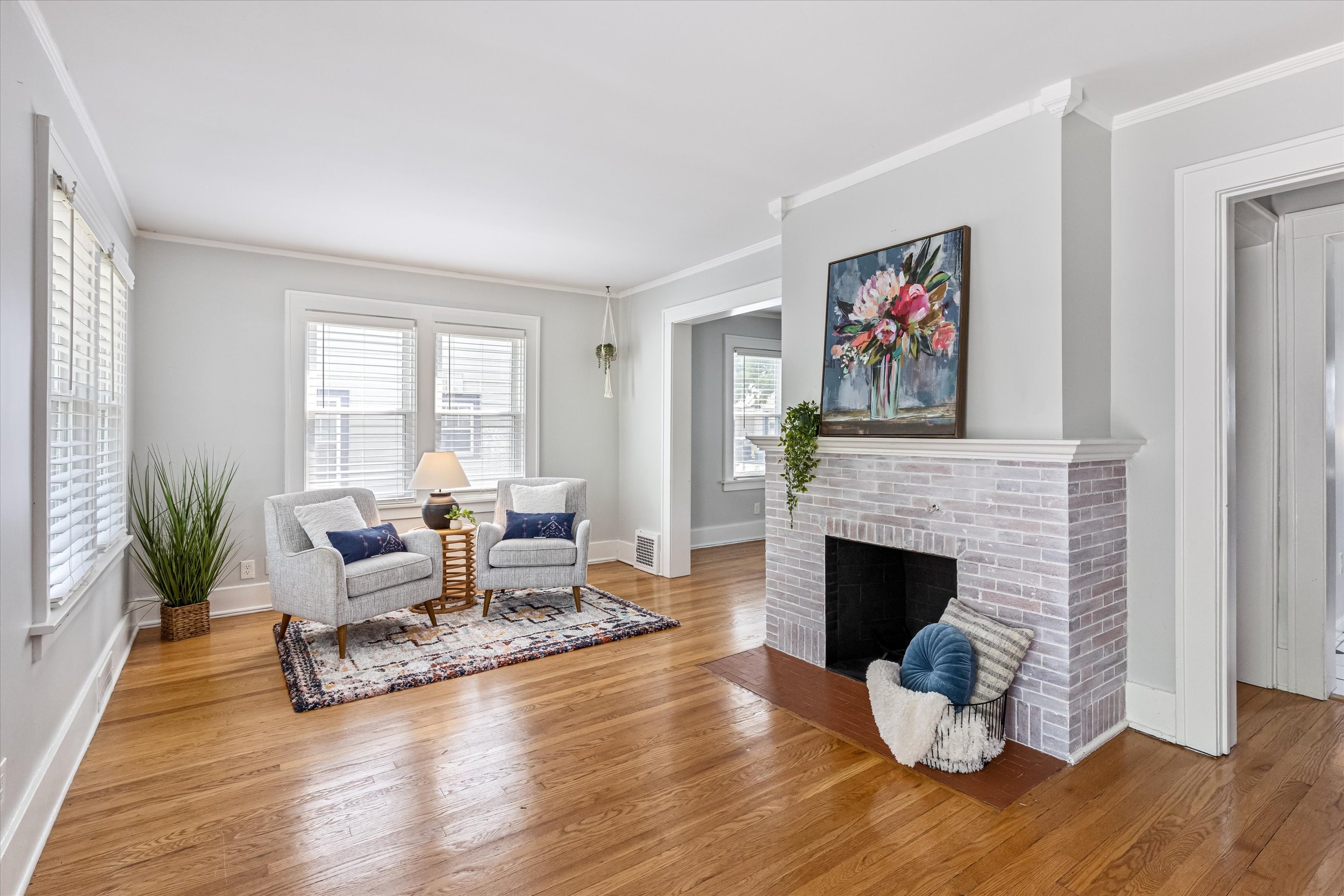 Bright living room with hardwood floors, white walls, and large windows with white blinds. There are two light gray armchairs with navy pillows in front of a window, a colorful patterned rug, and a fireplace with a painting of flowers above it. A bas