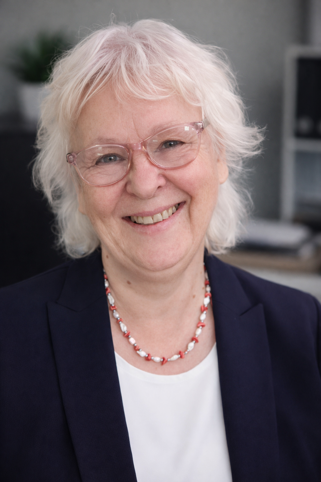 Smiling elderly woman with white hair and glasses, wearing a navy blazer and a red and white beaded necklace, in an office setting.