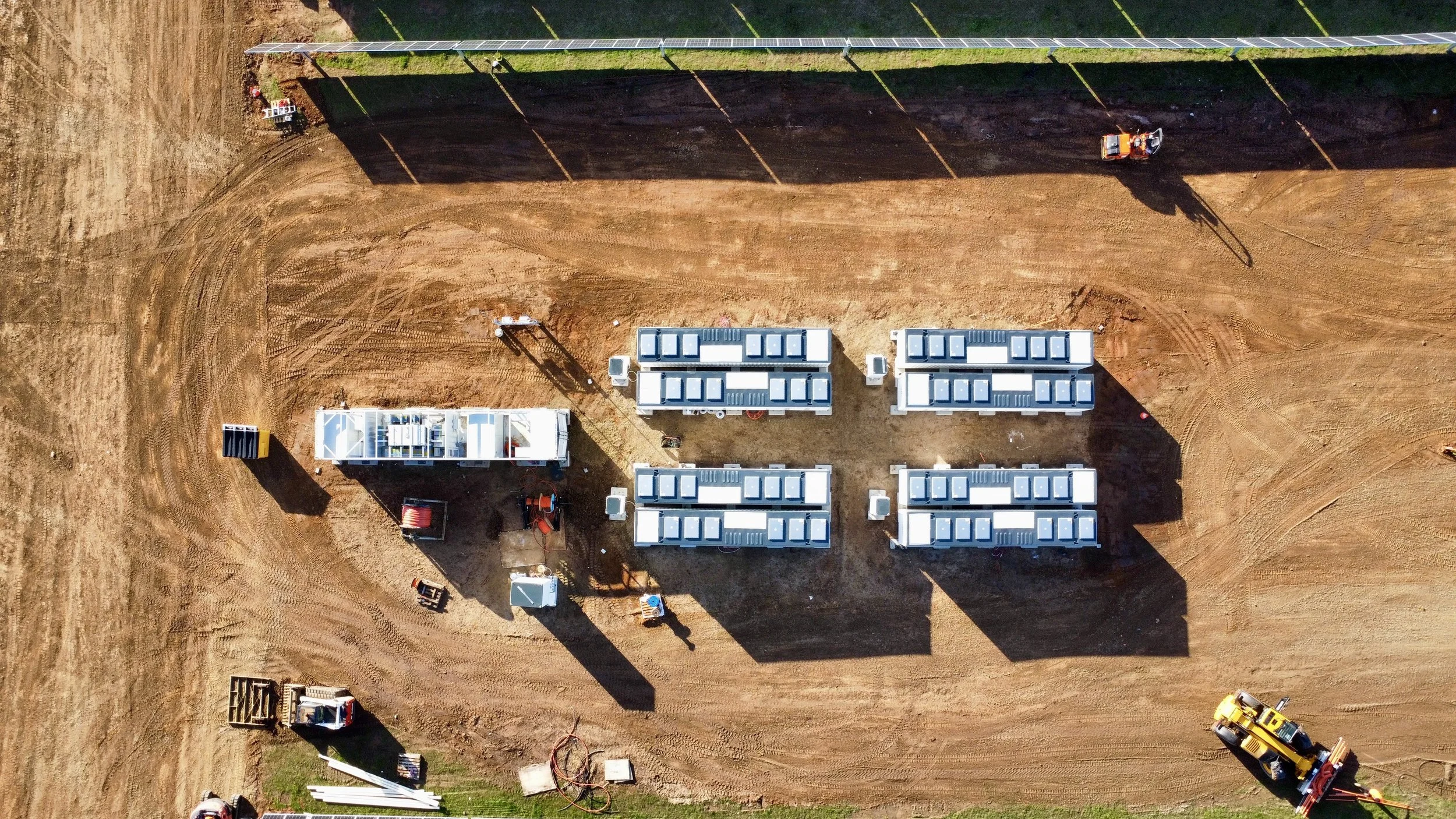 Aerial view of a construction site with several white modular units, a yellow excavator, and a roller. There is a dirt path, some construction equipment, and a solar panel array on a narrow strip of ground.