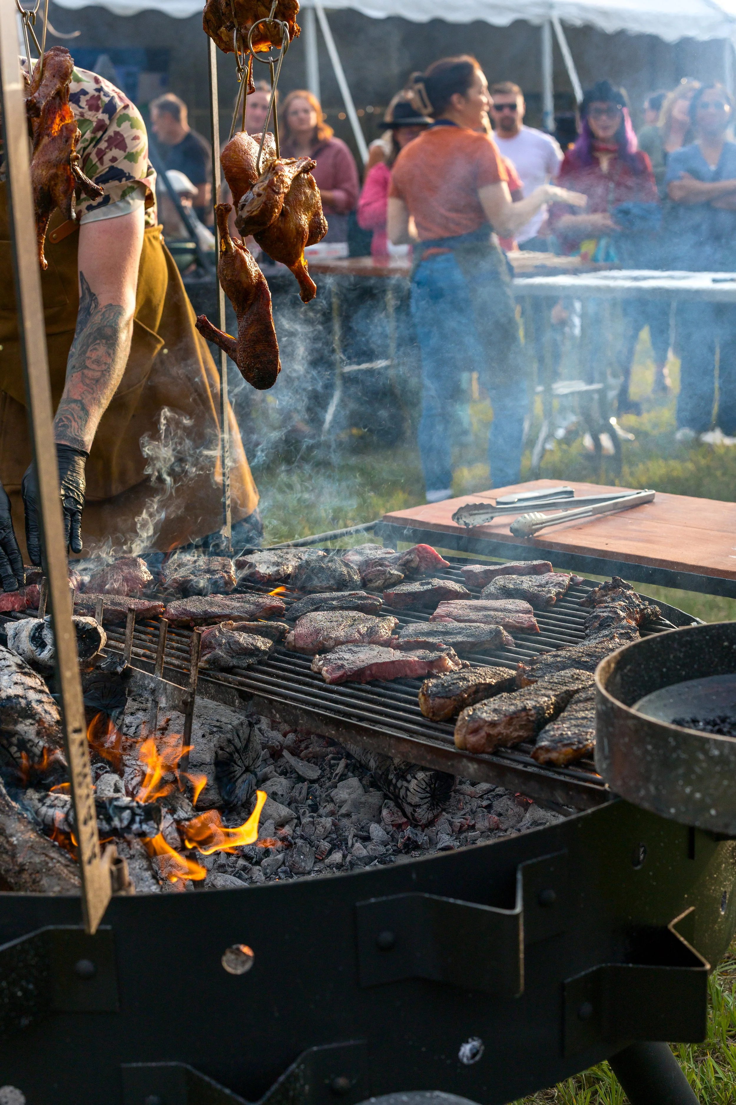 Barbecue grill with meat cooking over hot coals, hanging chicken, and people in background at an outdoor gathering.
