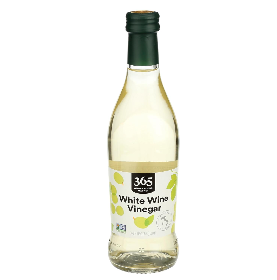 A clear glass bottle of 365 Whole Foods Market white wine vinegar with a green cap and a label featuring yellow grapes and a lemon, on a white background.