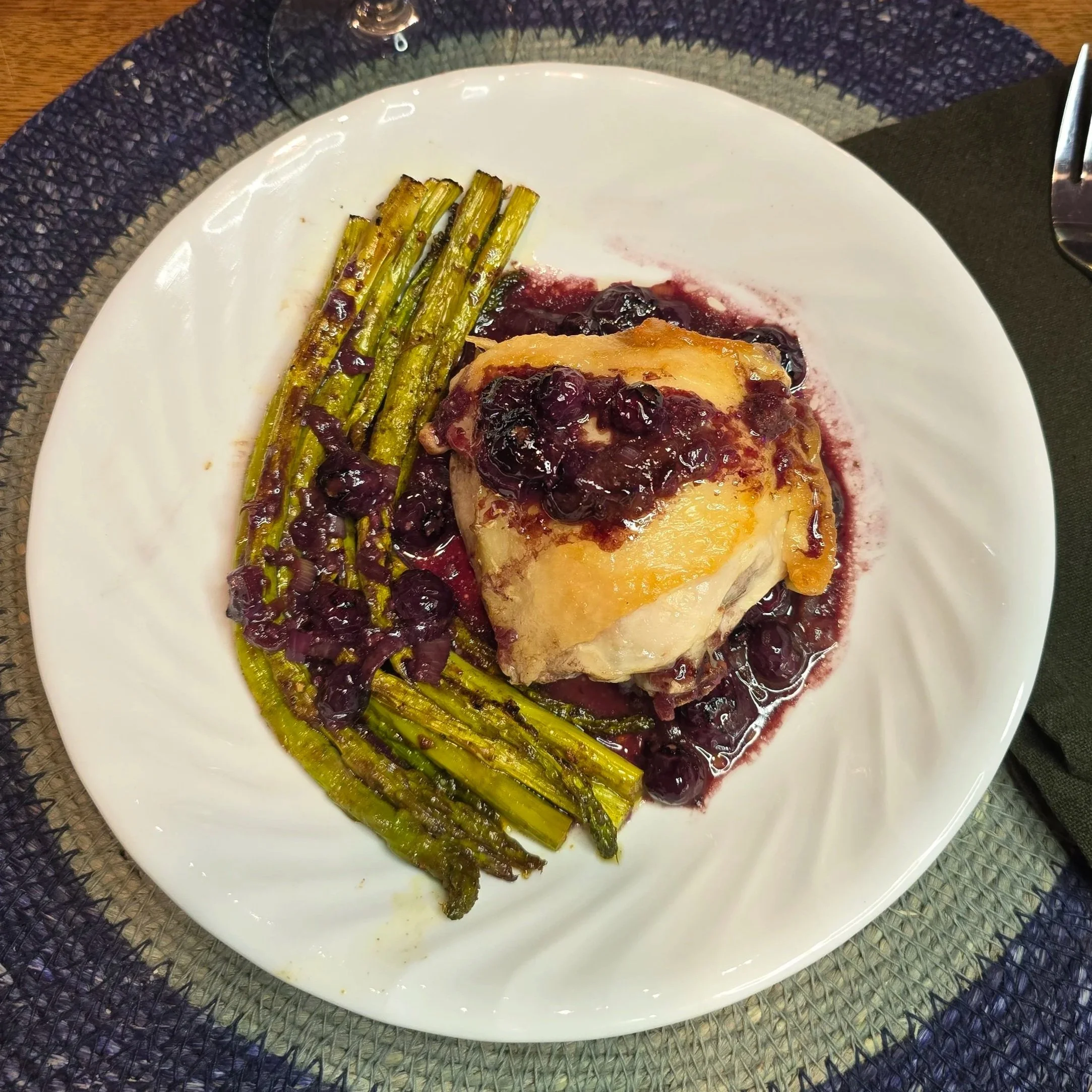 Roasted chicken thighs in a blueberry mustard reduced sauce with asparagus in a white ceramic plate on a blue mat.