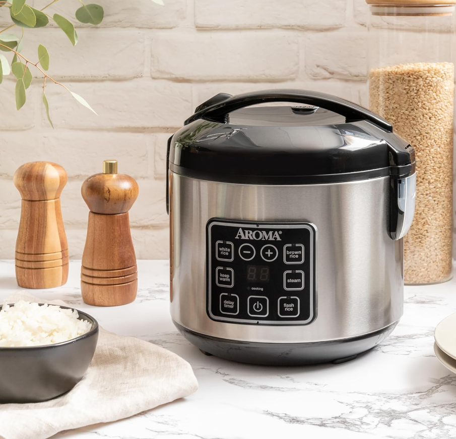 A stainless steel rice cooker on a white marble countertop with a black lid and control panel, next to a bowl of cooked rice and containers of rice and seasonings in a kitchen setting.