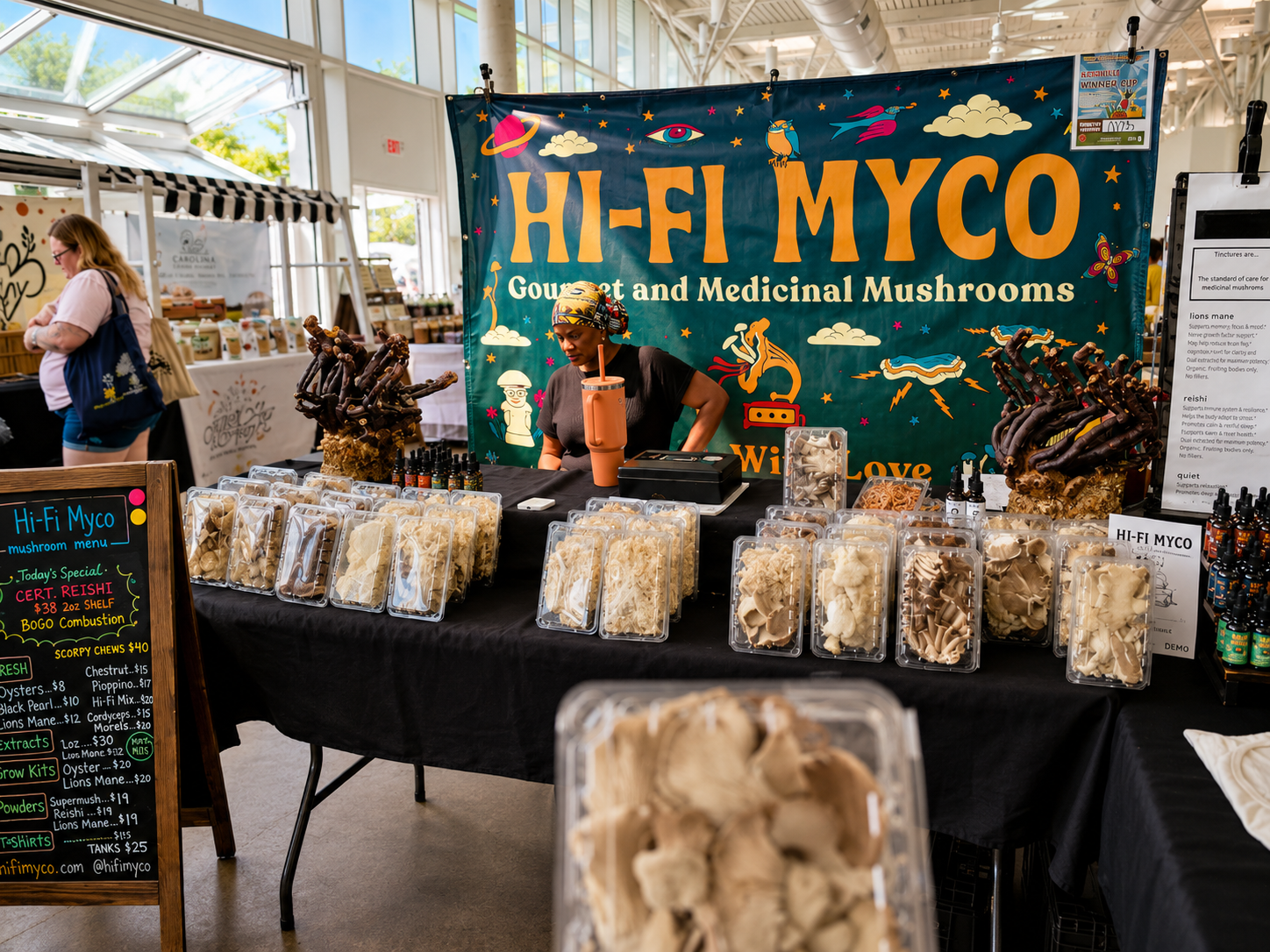 Hi-Fi Myco mushroom vendor stand at an indoor farmers market, featuring packages of fresh gourmet mushrooms, mushroom products, and a colorful vendor banner.