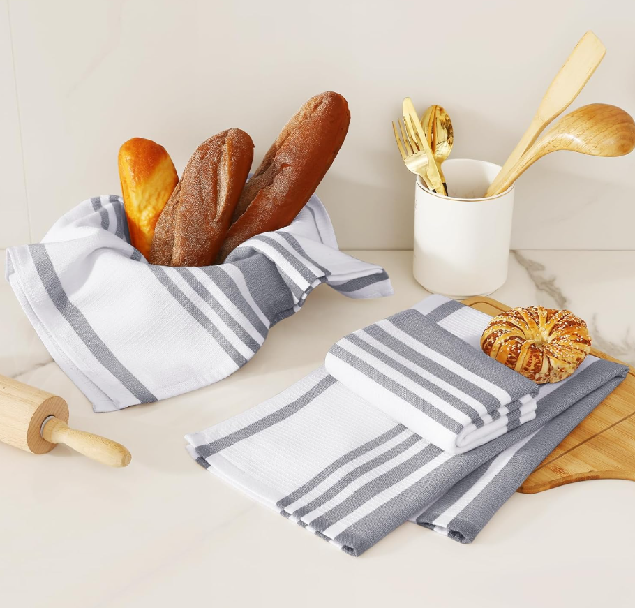 Fresh baked baguette bread in a striped cloth basket, a croissant on folded striped cloth napkins, and kitchen utensils in a white container, on a white marble countertop.