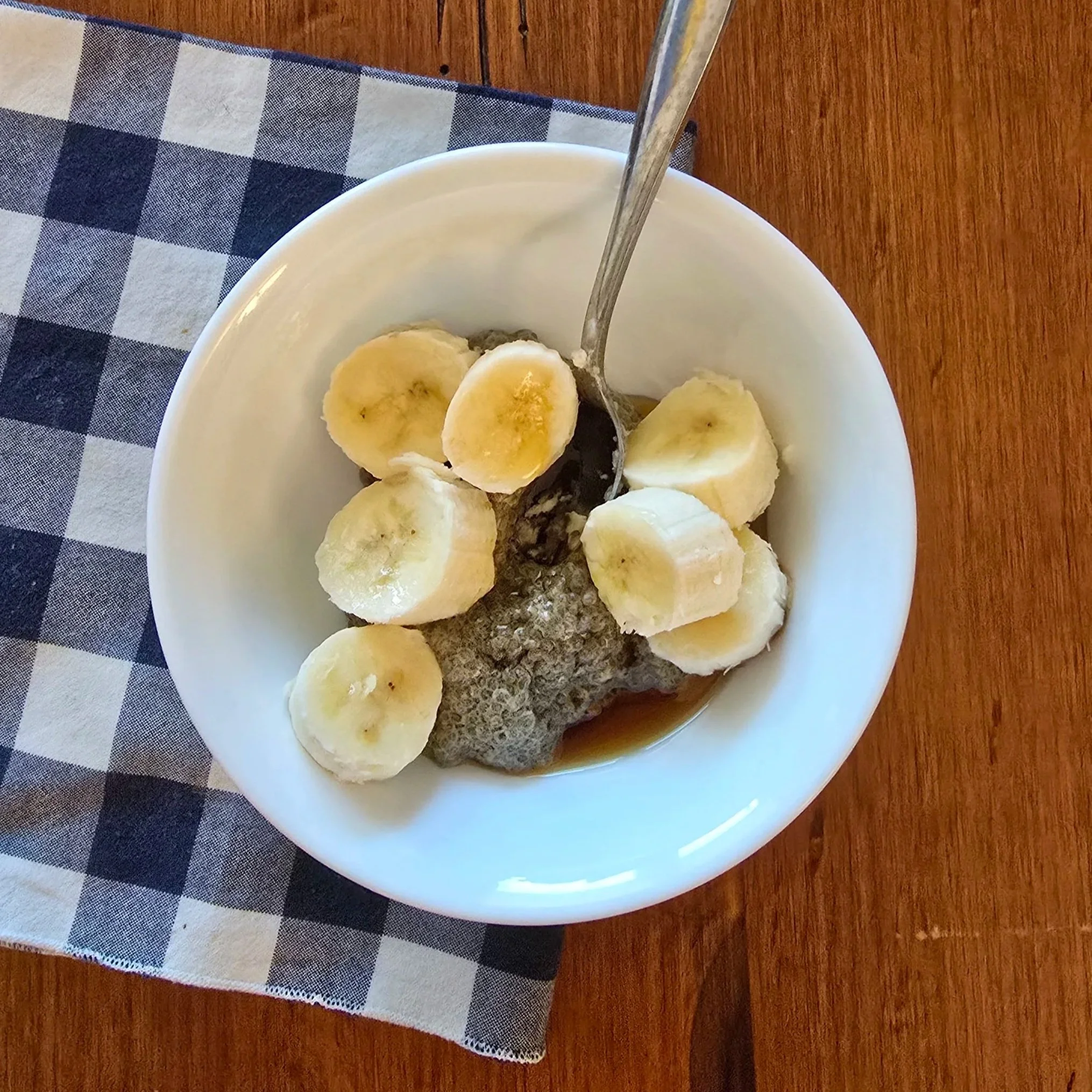 chia seed pudding with banana slices, maple syrup, and cinnamon in a white bowl with checkered napkin on a wood table