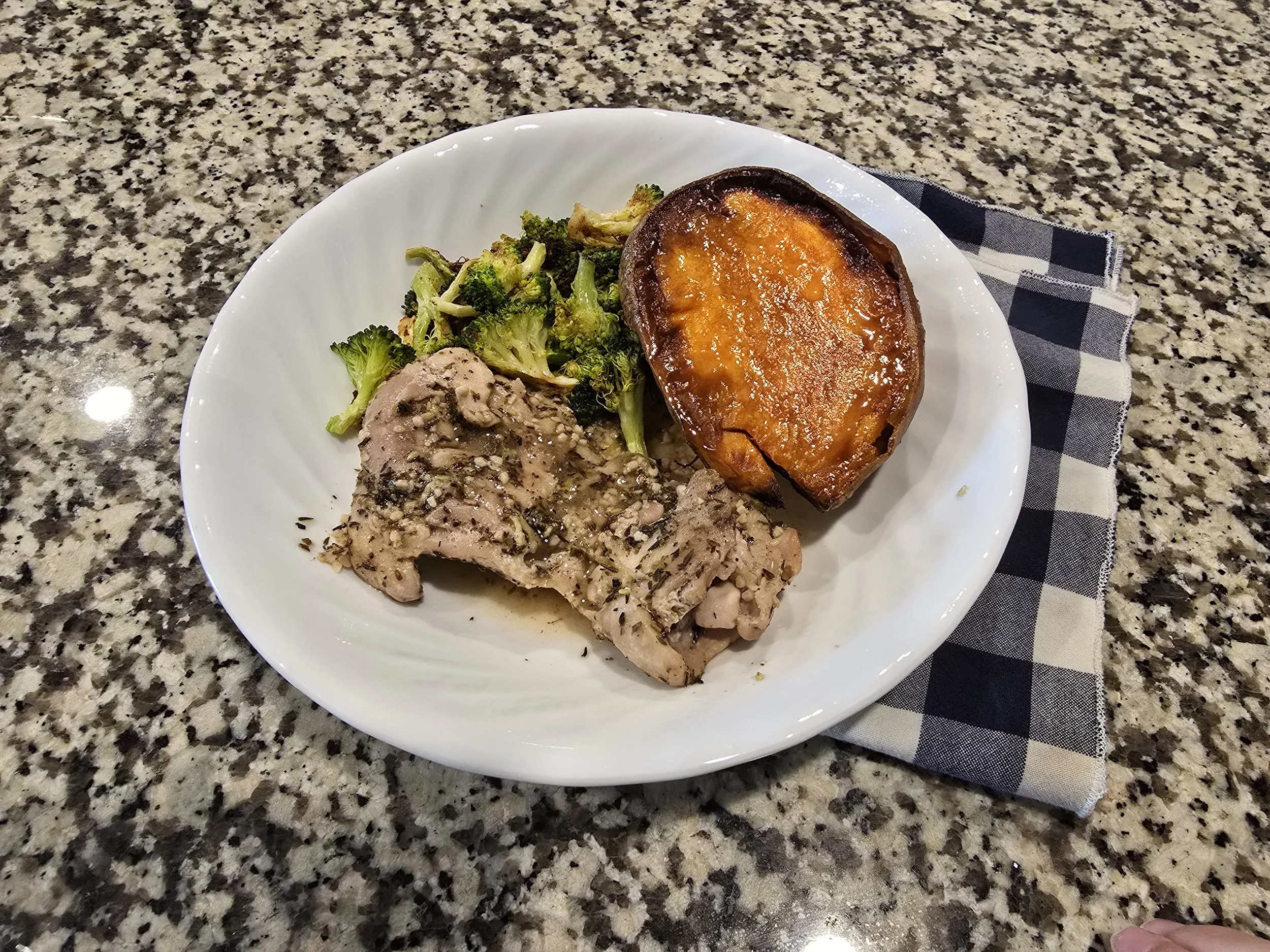 Roasted chicken thighs and baked sweet potatoes with air fried broccoli on a white plate with a cloth napkin on a granite counter top.
