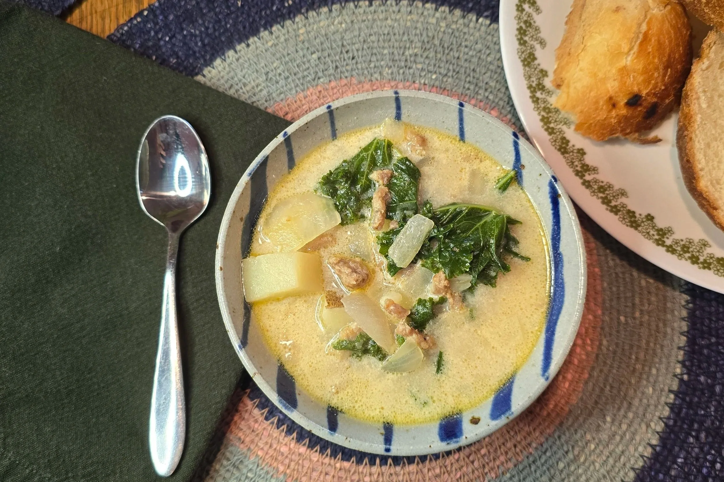 Copycat Zuppa Toscana Soup in a ceramic bowl with a spoon and cotton cloth napkin next to a vintage Correl green flower patterned plate of sourdough bread.