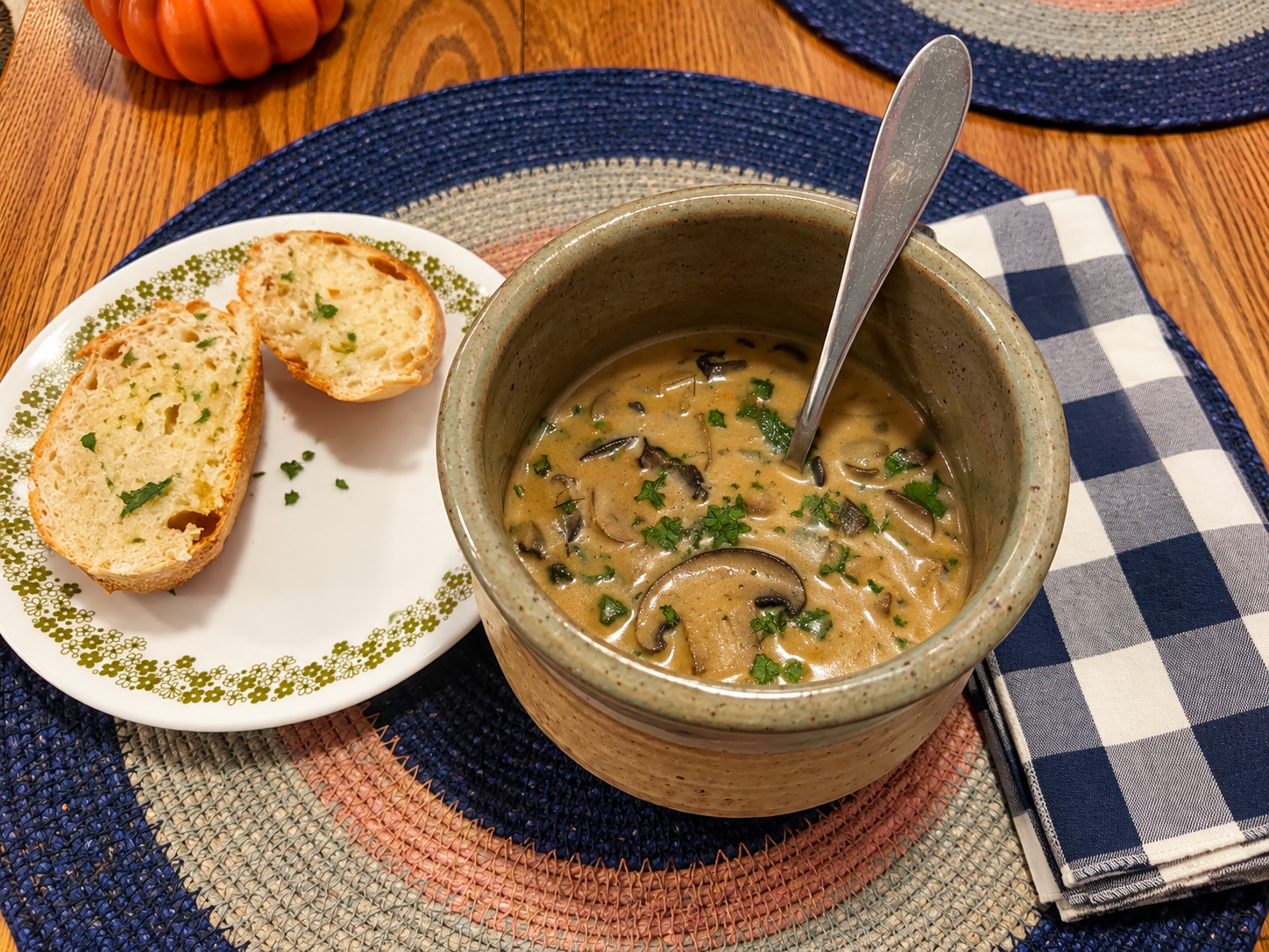 Creamy mushroom soup in a ceramic bowl with buttered sourdough bread on a wood table with blue and white checkered cotton napkin on a placemat.