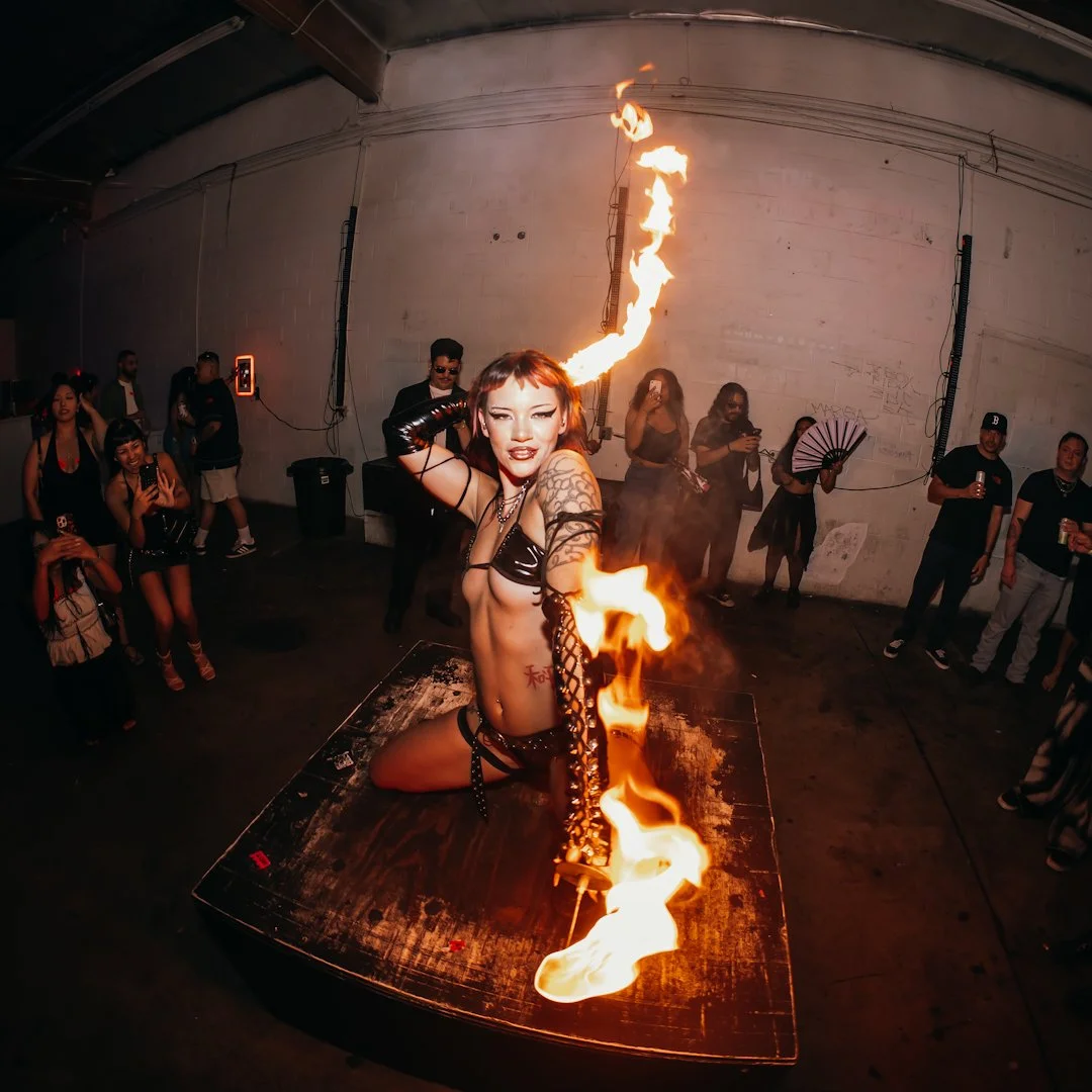 A fire performer with short hair, tattoos, and a black outfit is kneeling on stage, performing fire breathing at an indoor event with an audience watching.