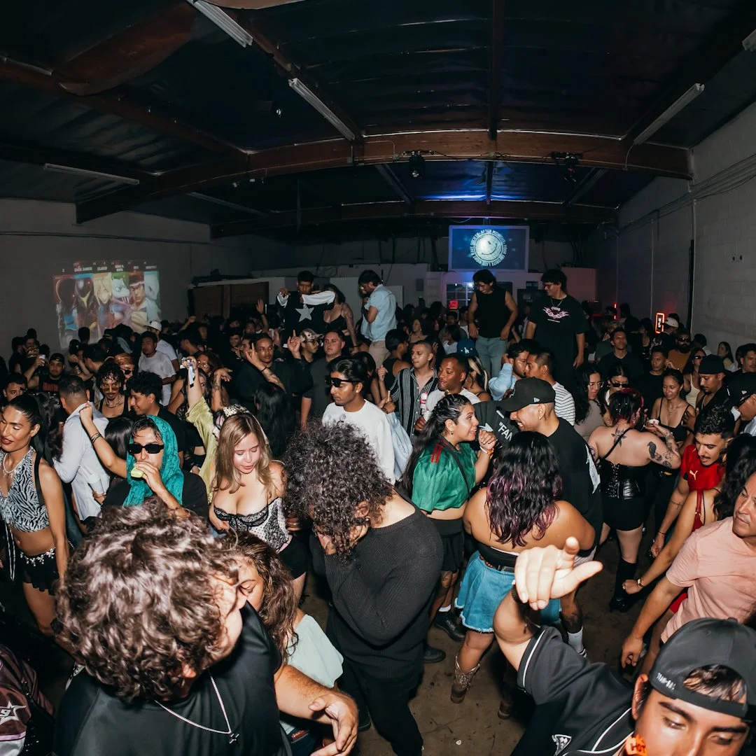 Crowded indoor dance party with diverse people dancing and socializing, a large screen on the back wall displaying a logo, and colorful lighting.