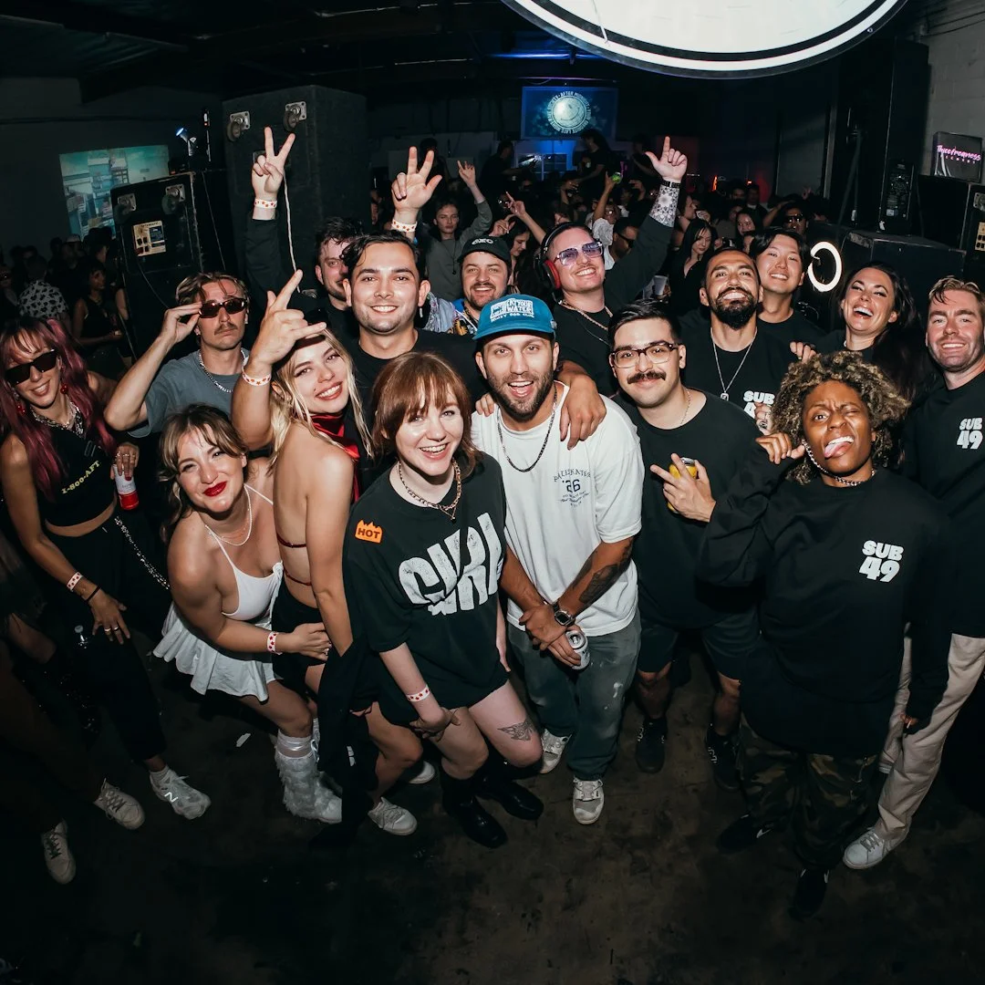 Group of smiling people at a party or concert, posing for a photo with some making peace signs, in an indoor venue with stage lights and a crowd in the background.