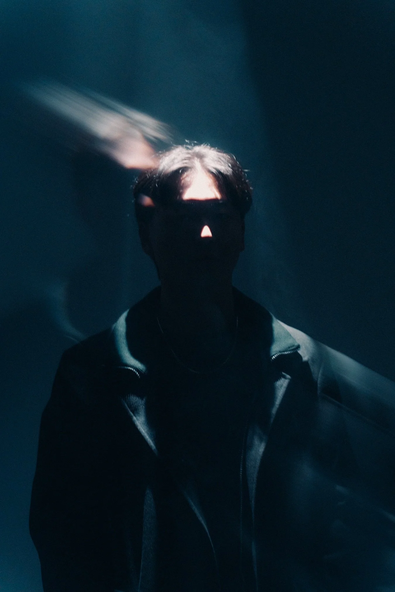a person with dark hair wearing a black face mask and a dark jacket, with a dark background and light streaks near their head
