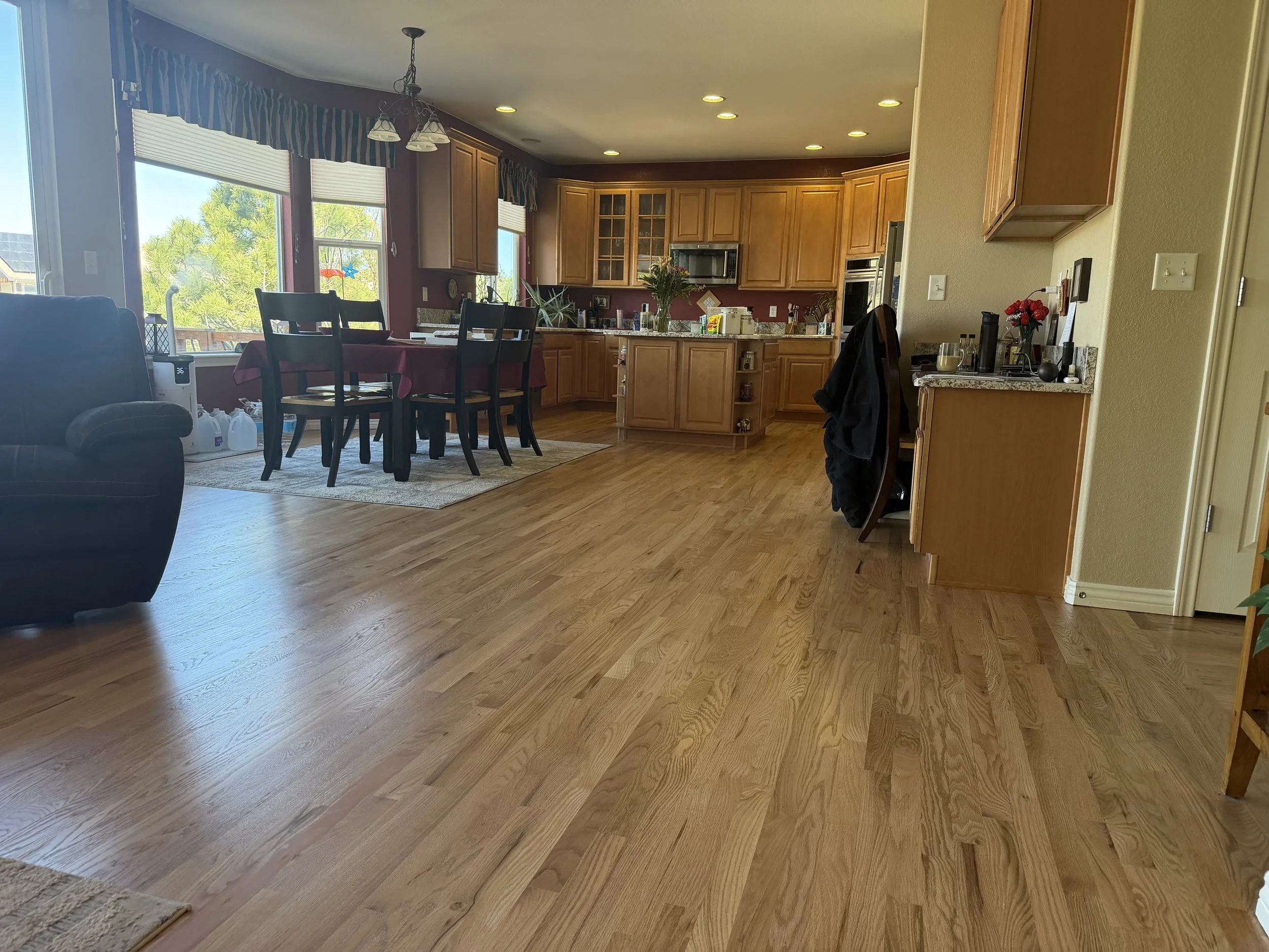 view of a hardwood floor project in Castle Rock/Castle Pines, Colorado with light hardwood floors spanning across an open-floor plan spanning the living space, dining space, and kitchen. The kitchen has light cabinets and an island in the middle.