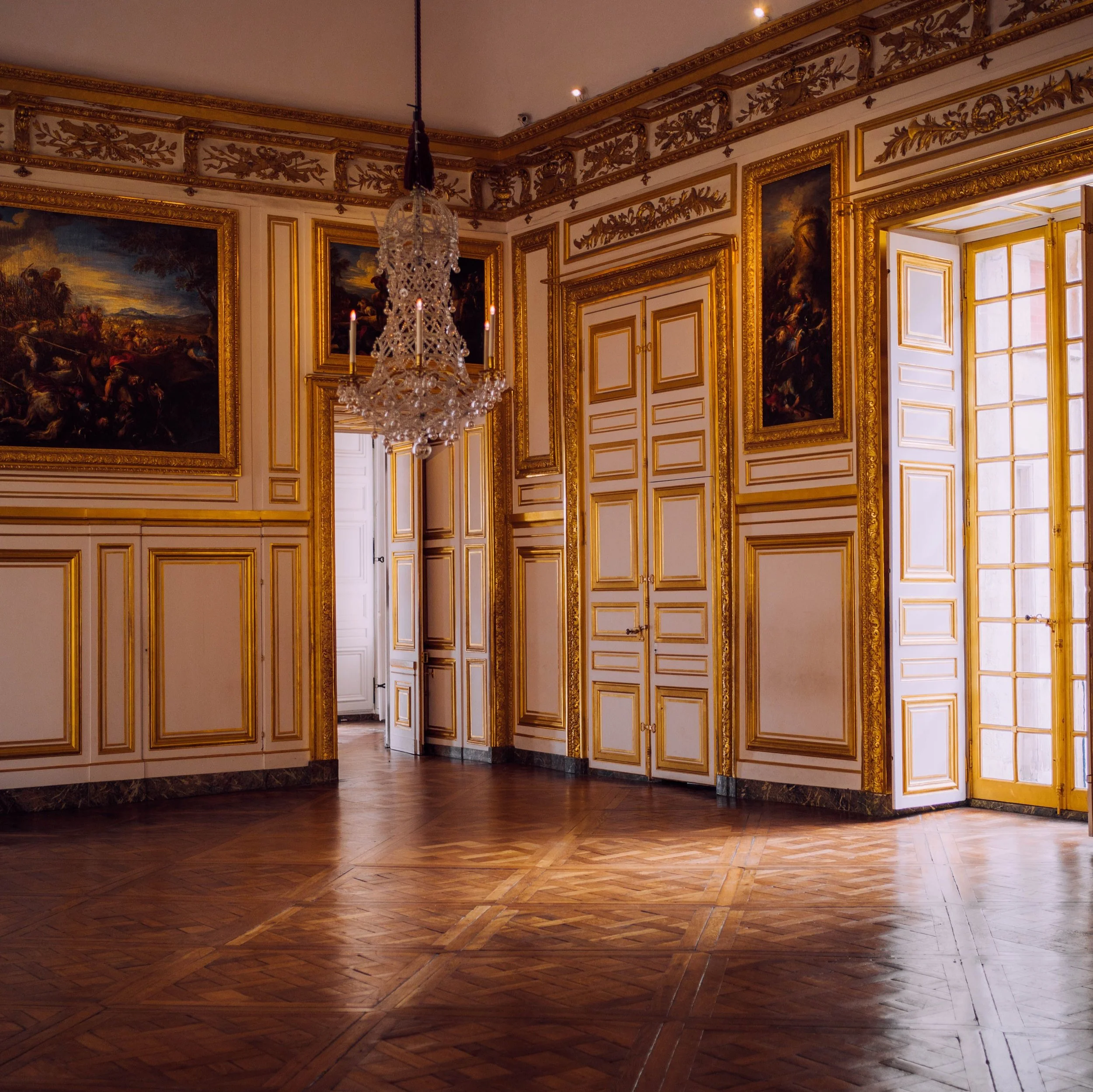 view of a room within the Grand Trianon in Versailles with beautiful, ornate walls containing gold accents and large oil paintings. There is a chandellier and a beautiful hardwood floor with an intricate design.