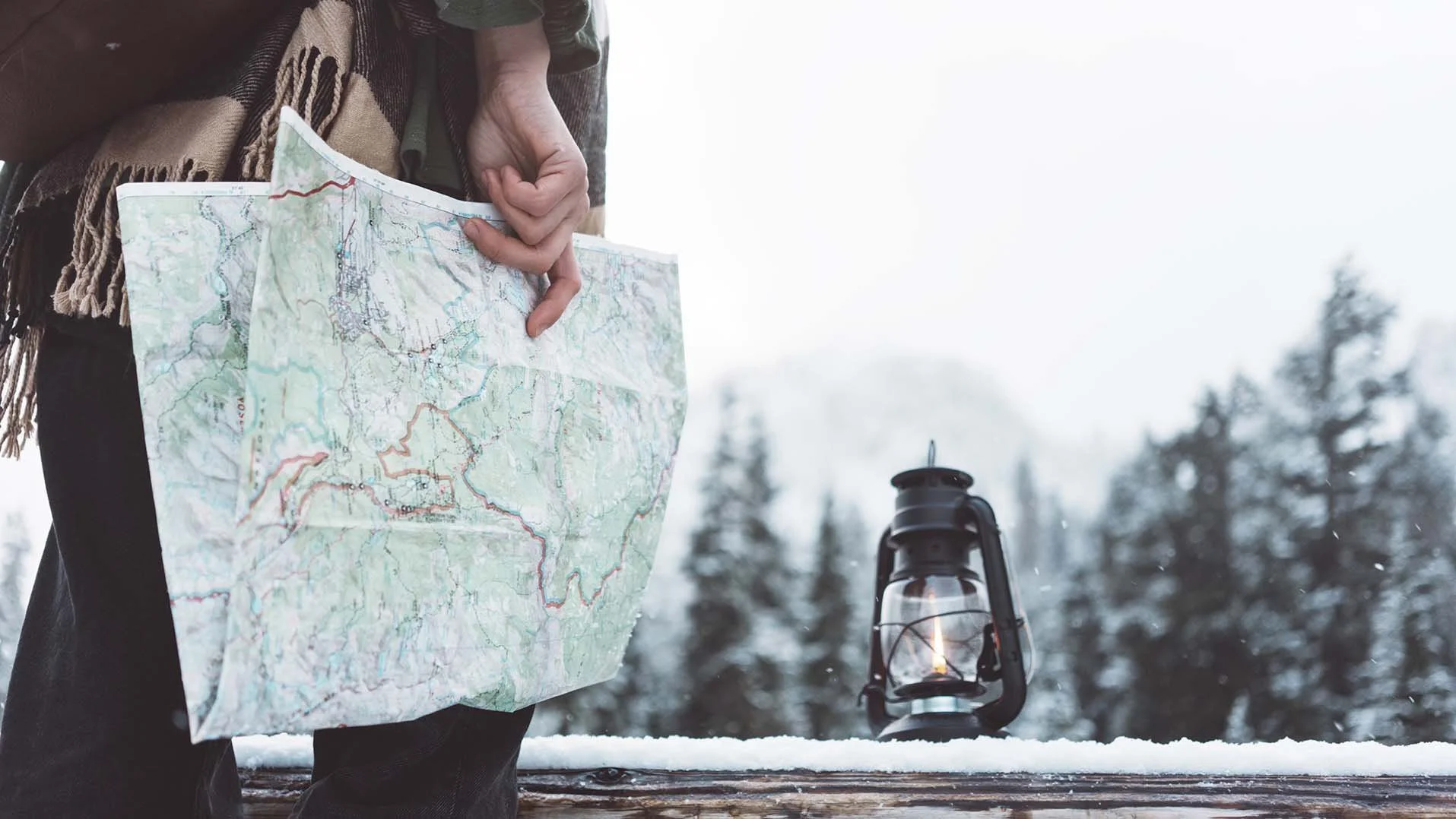 Winter hiker holding a Mt. Hood trail map beside a lantern on a snowy deck near RAVN HAUS modern alpine vacation rental in Government Camp, Oregon.