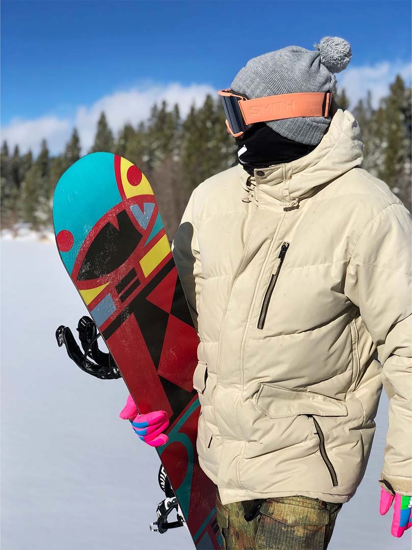 Snowboarder in a white jacket and goggles holding a colorful graphic snowboard on a bluebird powder day at Mt. Hood near RAVN HAUS modern alpine vacation rental in Government Camp, Oregon.