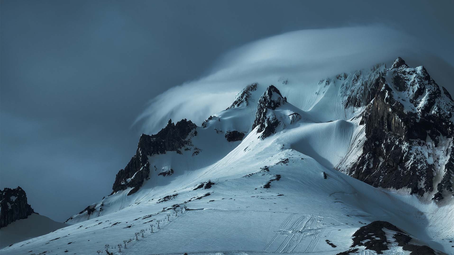Dramatic winter landscape of Mt. Hood near Government Camp, Oregon, with stormy skies, wind-sculpted snowfields, and ski tracks carving the lower slopes, inviting ski and snowboard adventures near RAVN HAUS alpine vacation rental.