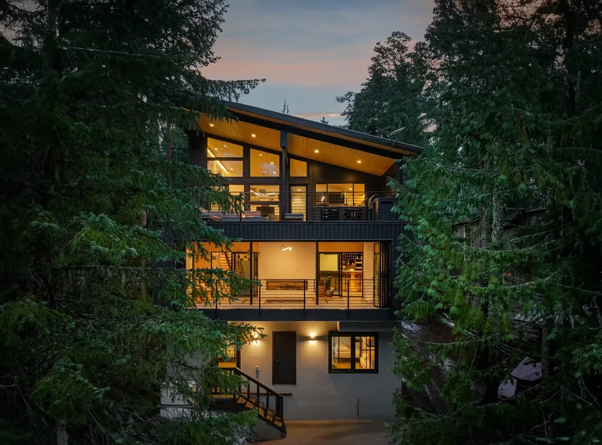 Illuminated rear view of RAVN HAUS Mt. Hood vacation rental showcasing the spa, patio seating, and tall windows nestled in the forest.