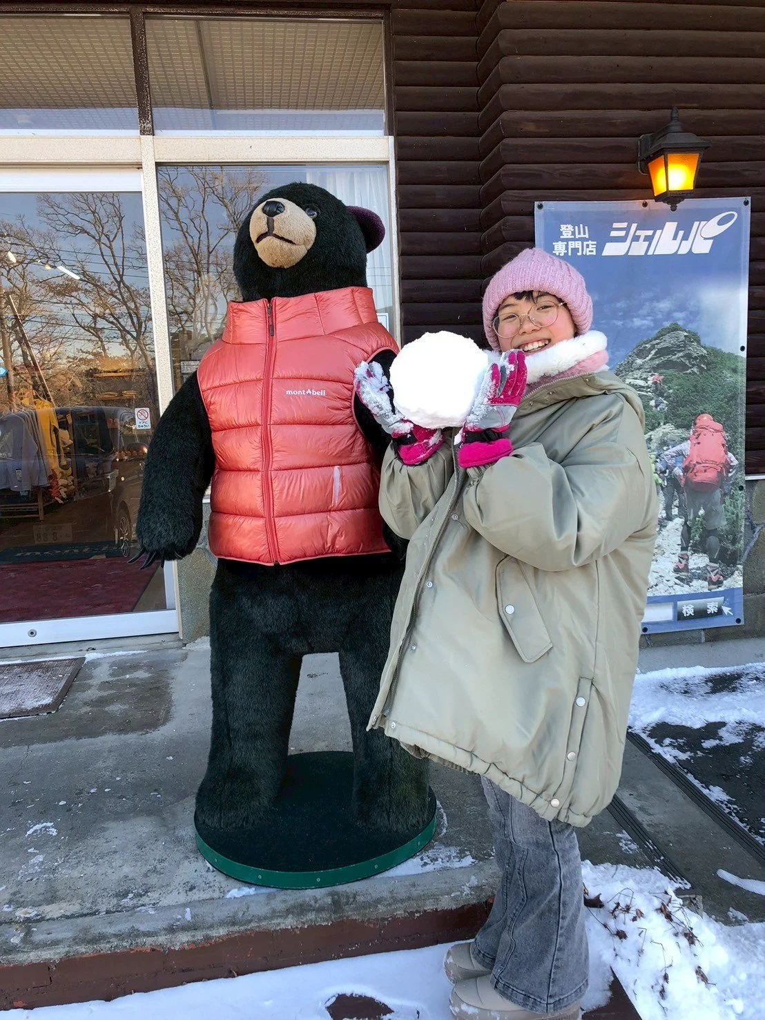 雪だるまを持って笑顔の女性と、クマのキャラクターのマスコットが登山店の前に立っている。背景には登山のポスターと建物の窓が見える。冬の景色で雪が積もっている。