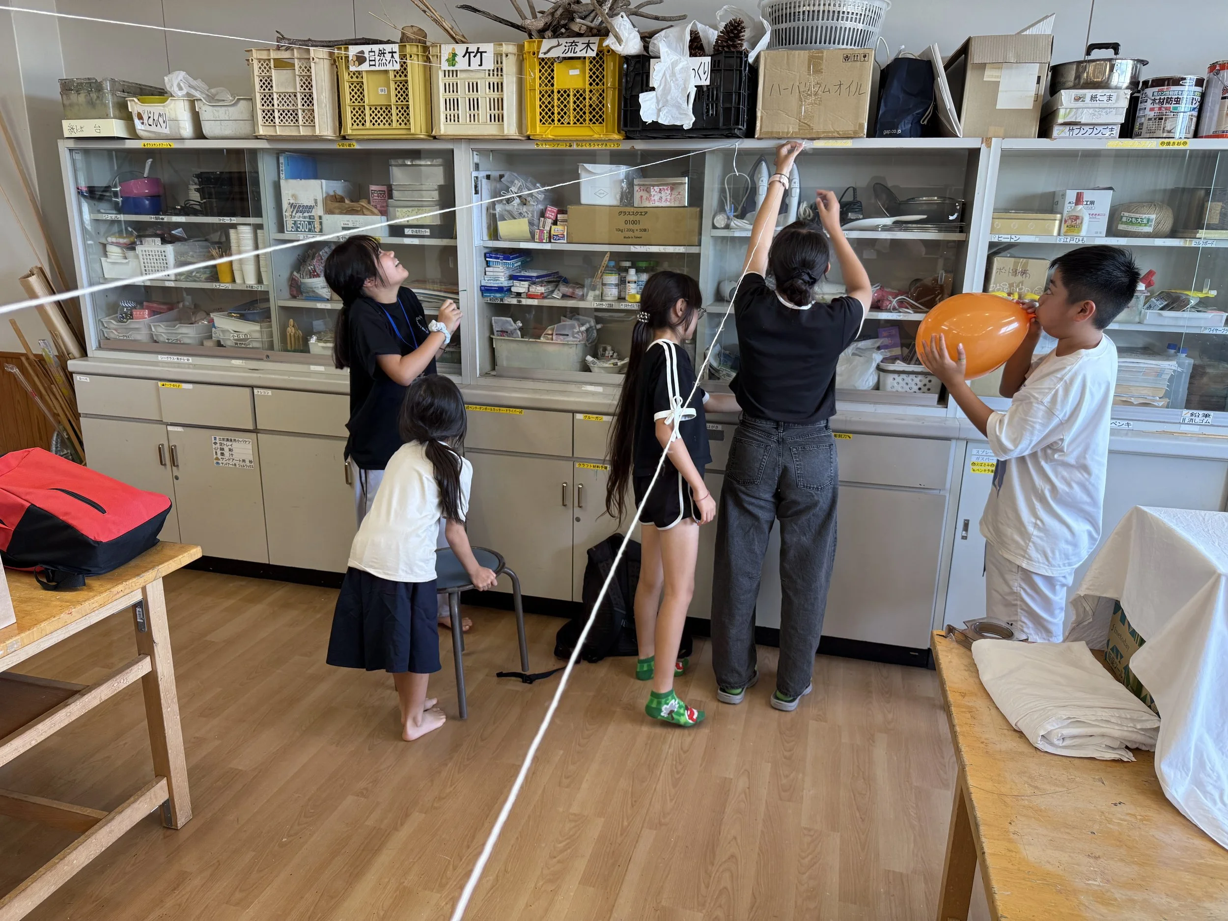 室内で子供たちが風船遊びをしている様子。中央の女の子が空に向かってジャンプし、他の子供たちが見守っている。背後には収納棚と様々な物品。